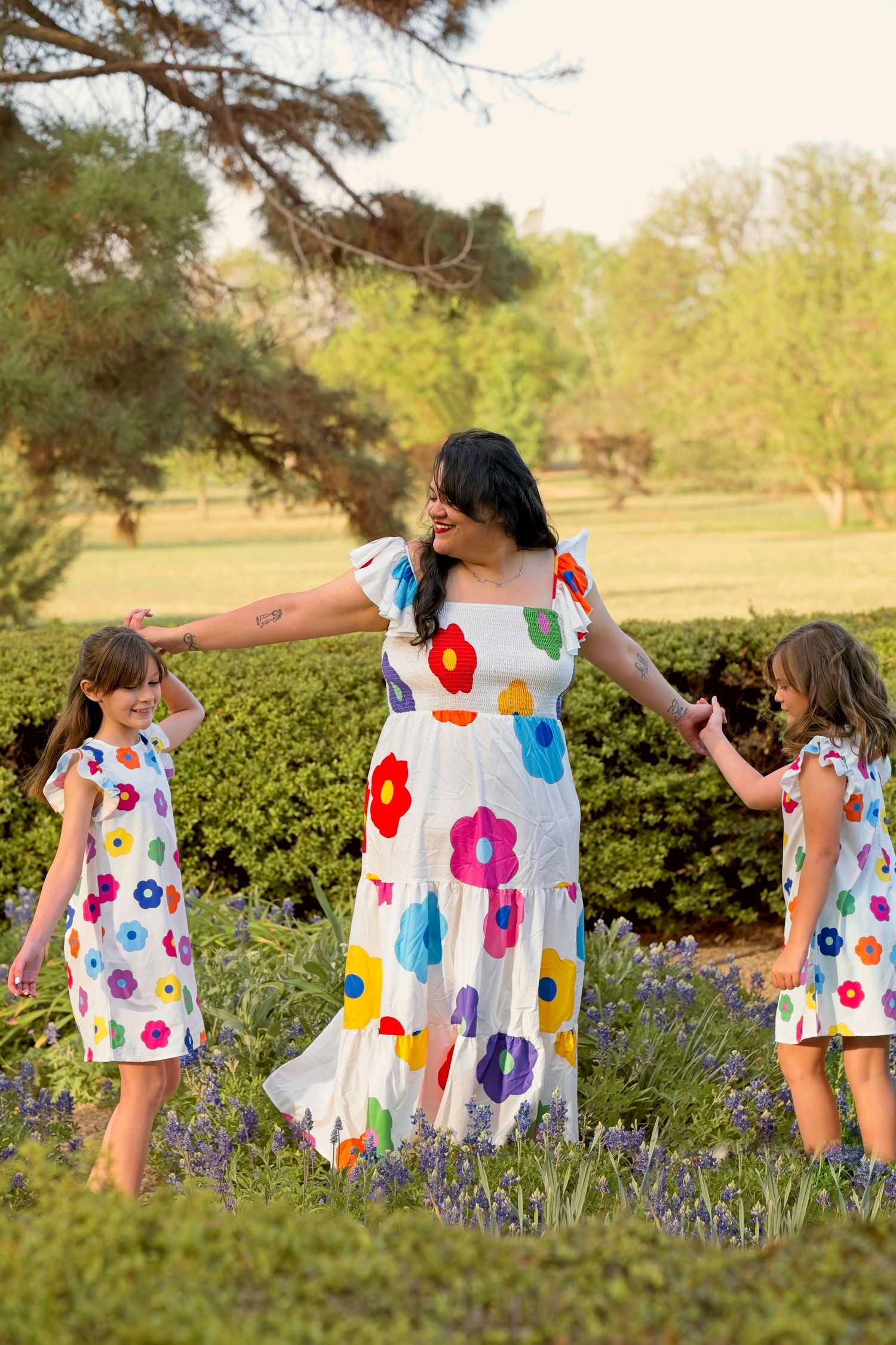 Three people in coordinating floral dresses hold hands and dance in a garden setting.