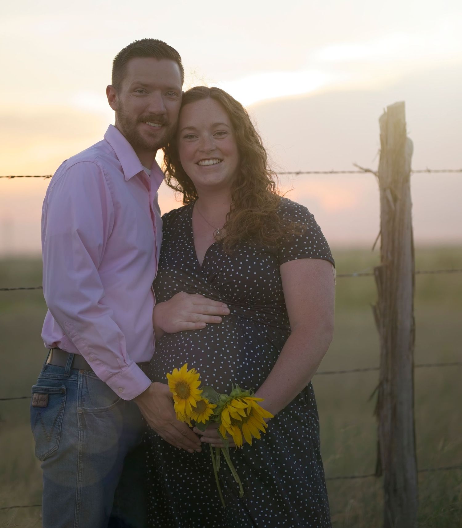 Couple embraces in a field with sunflowers at sunset while wearing pink shirt and polka dot dress.