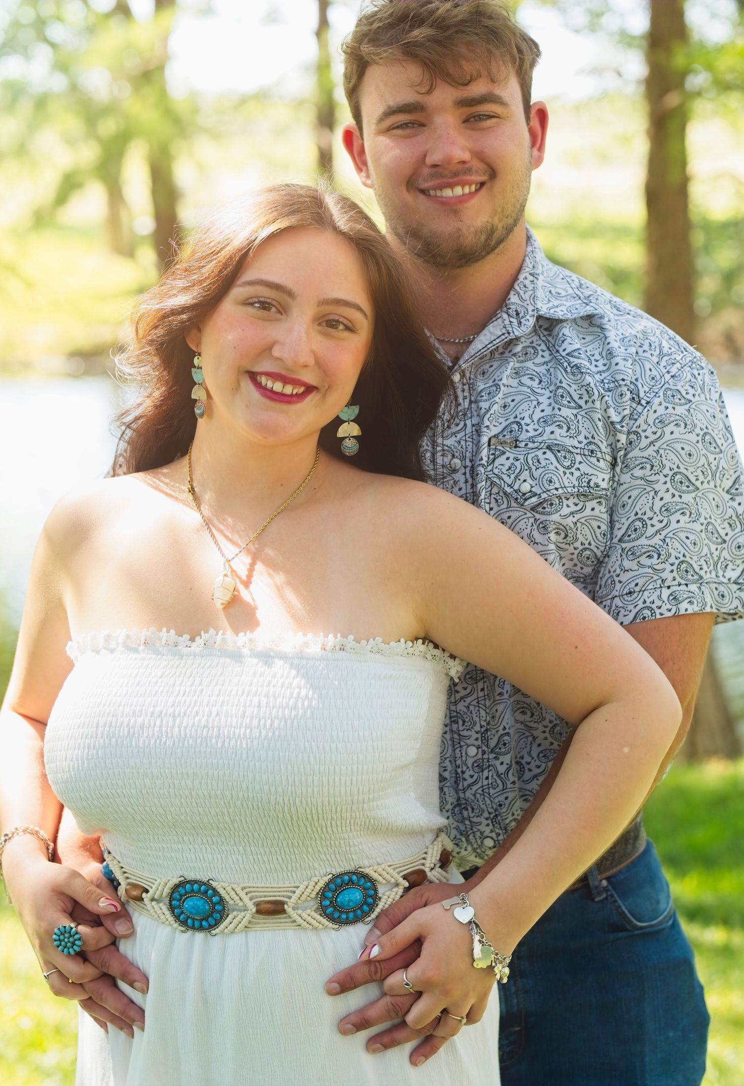 A couple poses together in nature on a sunny day wearing summer attire and sharing an intimate embrace.