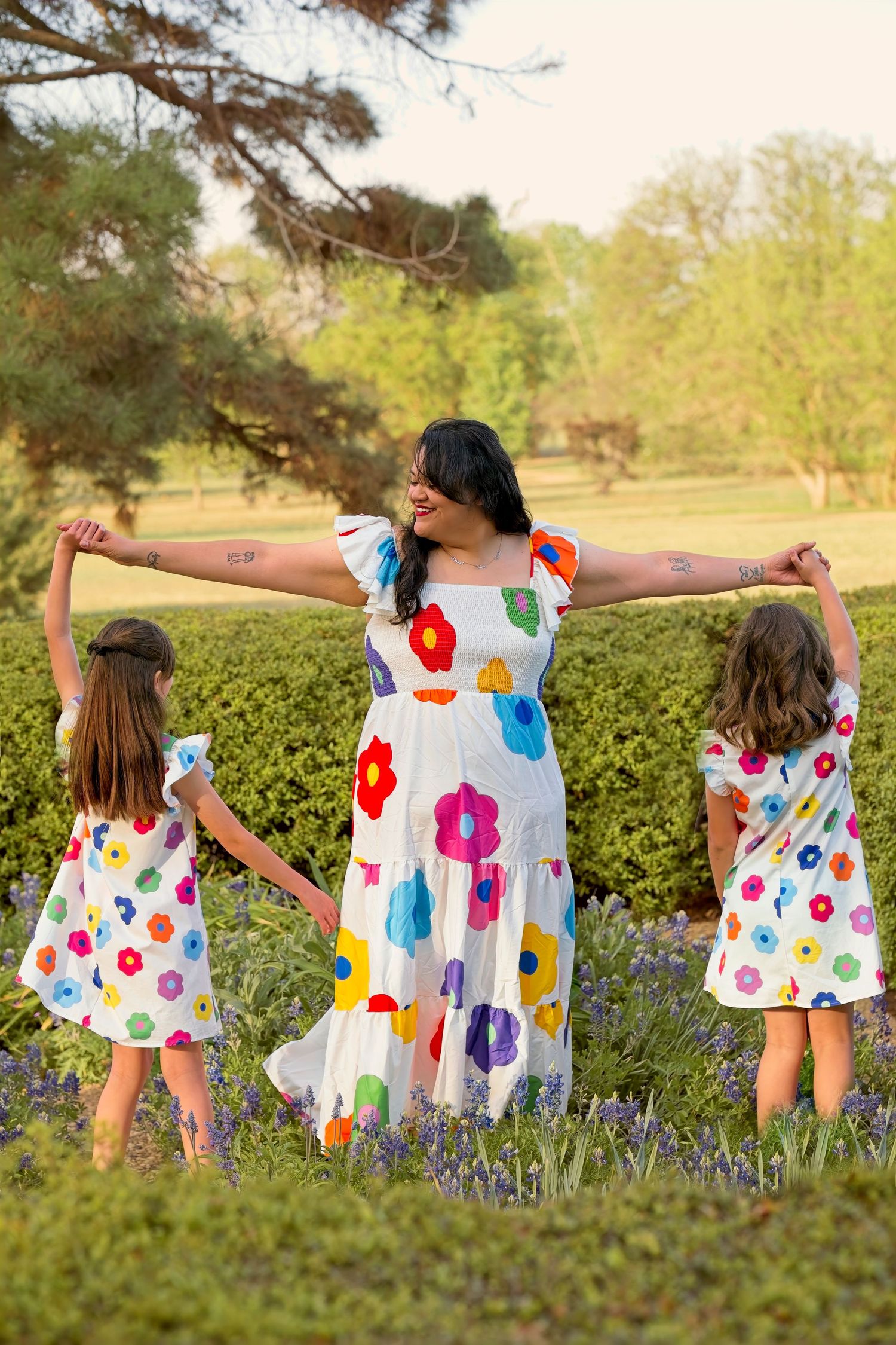 Three people in matching floral print dresses hold hands while standing in a blooming spring garden at sunset.