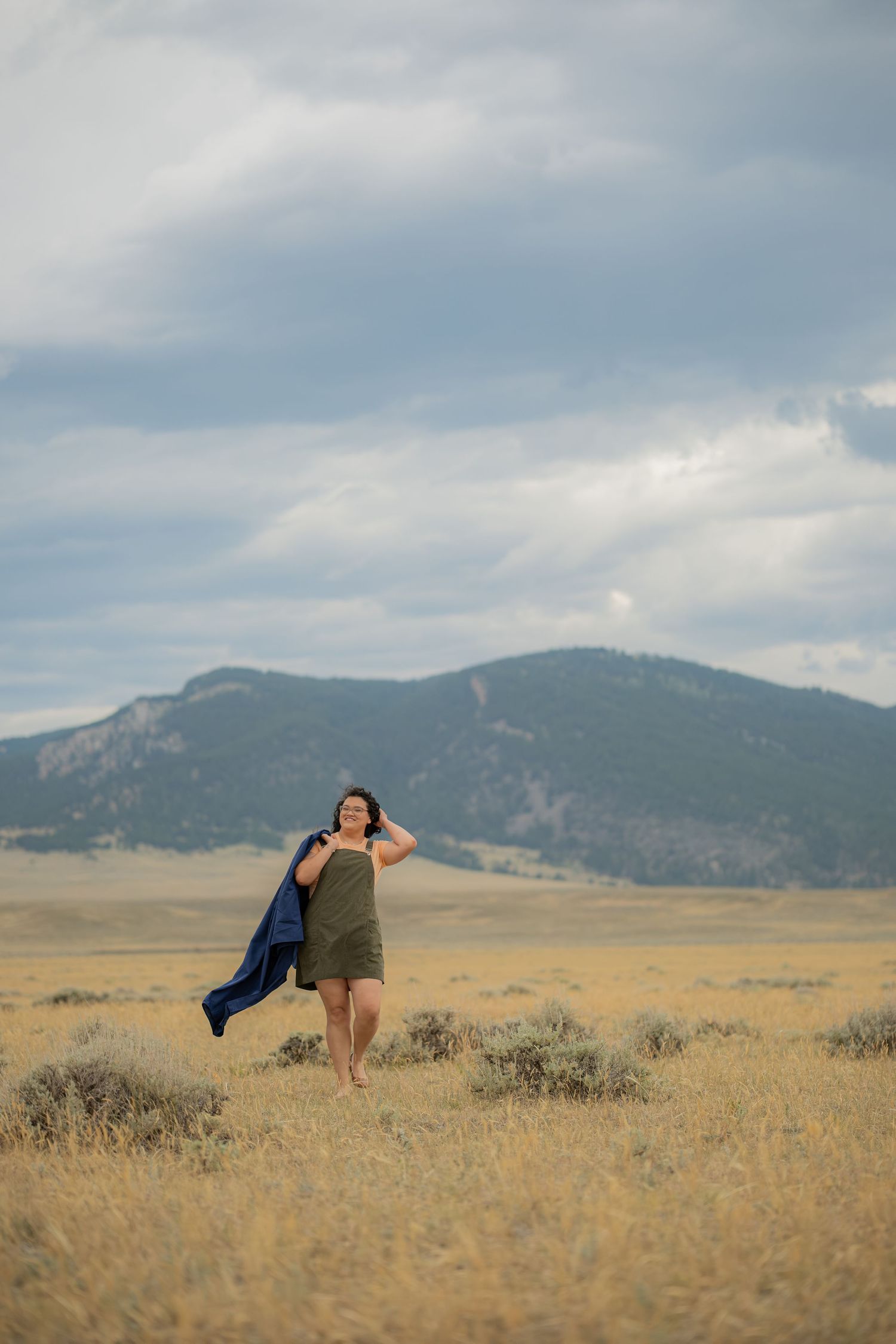 Woman in olive dress holding blue jacket stands barefoot in golden prairie field with mountain backdrop under cloudy sky.