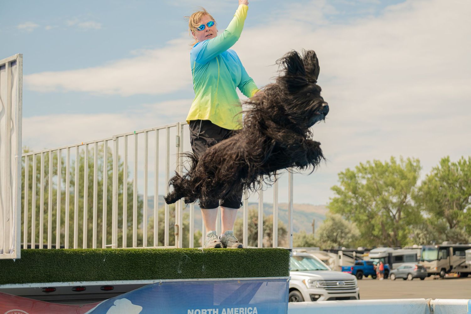 A large black fluffy dog leaps mid-air at a dock diving competition while a colorful handler cheers nearby.