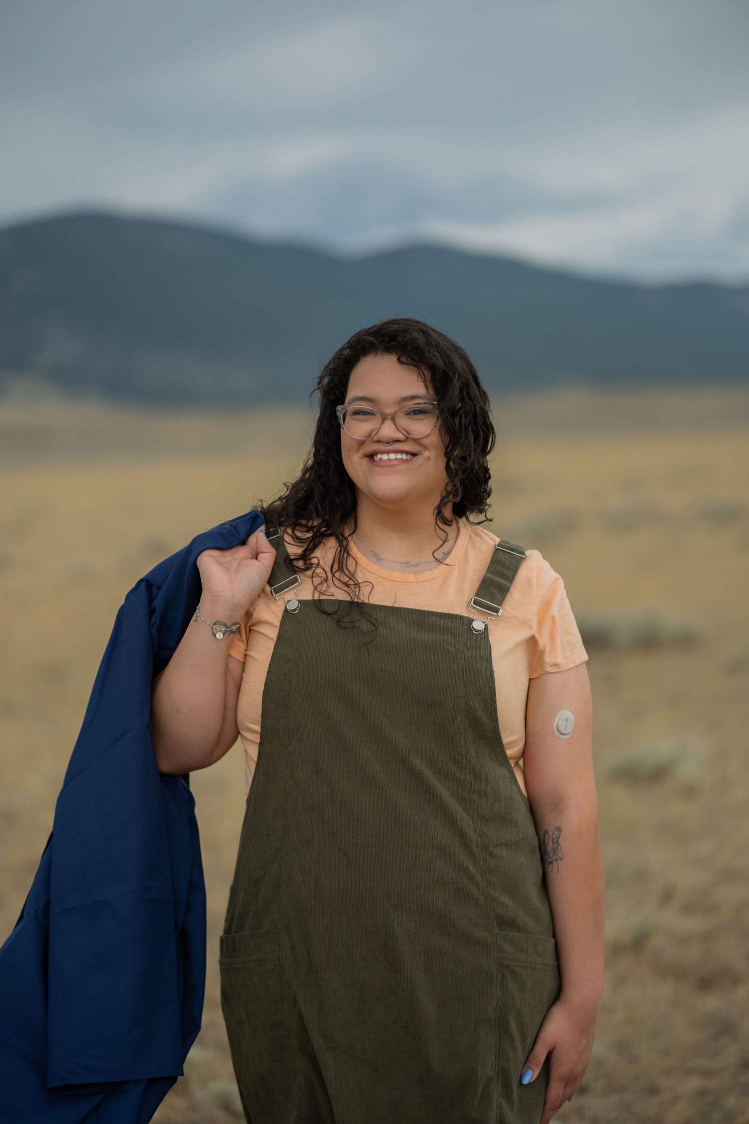 Smiling young woman in olive overalls and peach shirt holding blue jacket in open field with mountain backdrop.