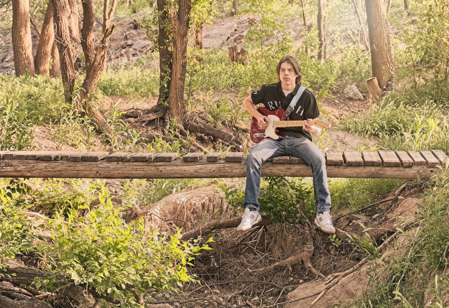 Guitarist performing while sitting on wooden bridge in wooded area.