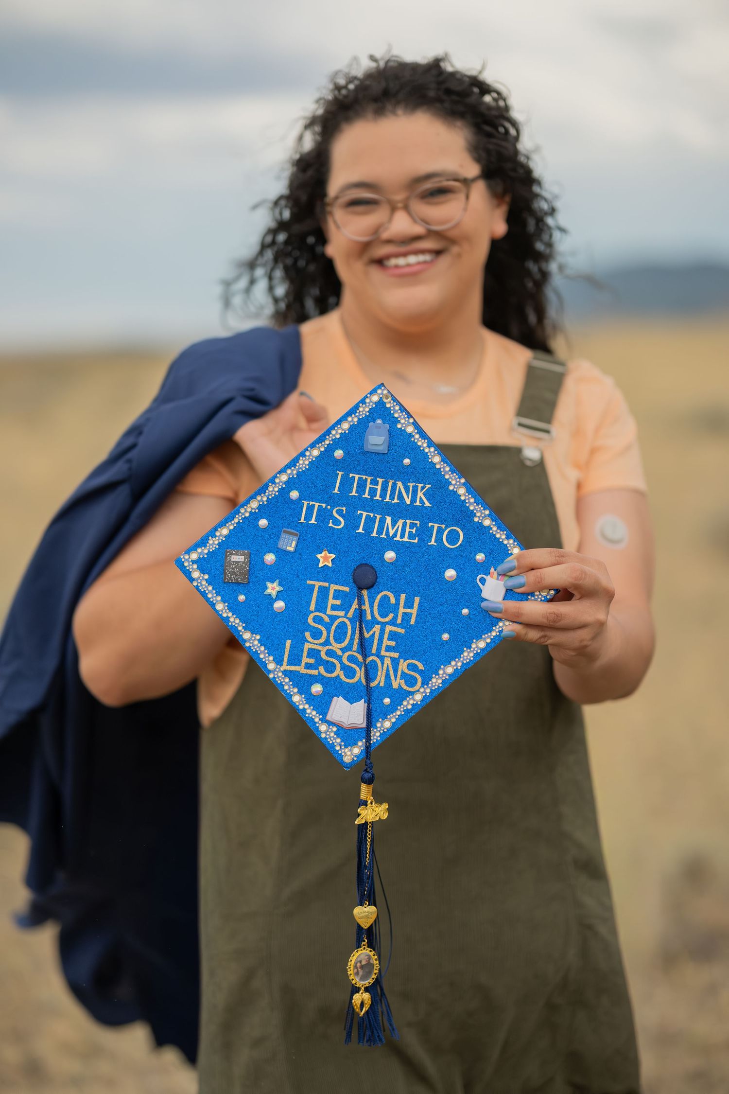 Smiling graduate holds blue decorated mortarboard cap reading I Think Its Time To Teach Some Lessons outdoors.
