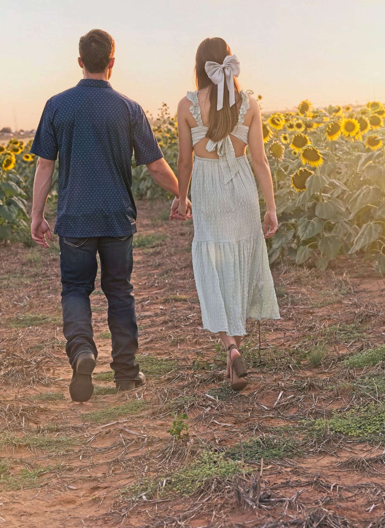 A couple walks hand in hand through a sunflower field at golden hour with long shadows and soft sunset light.