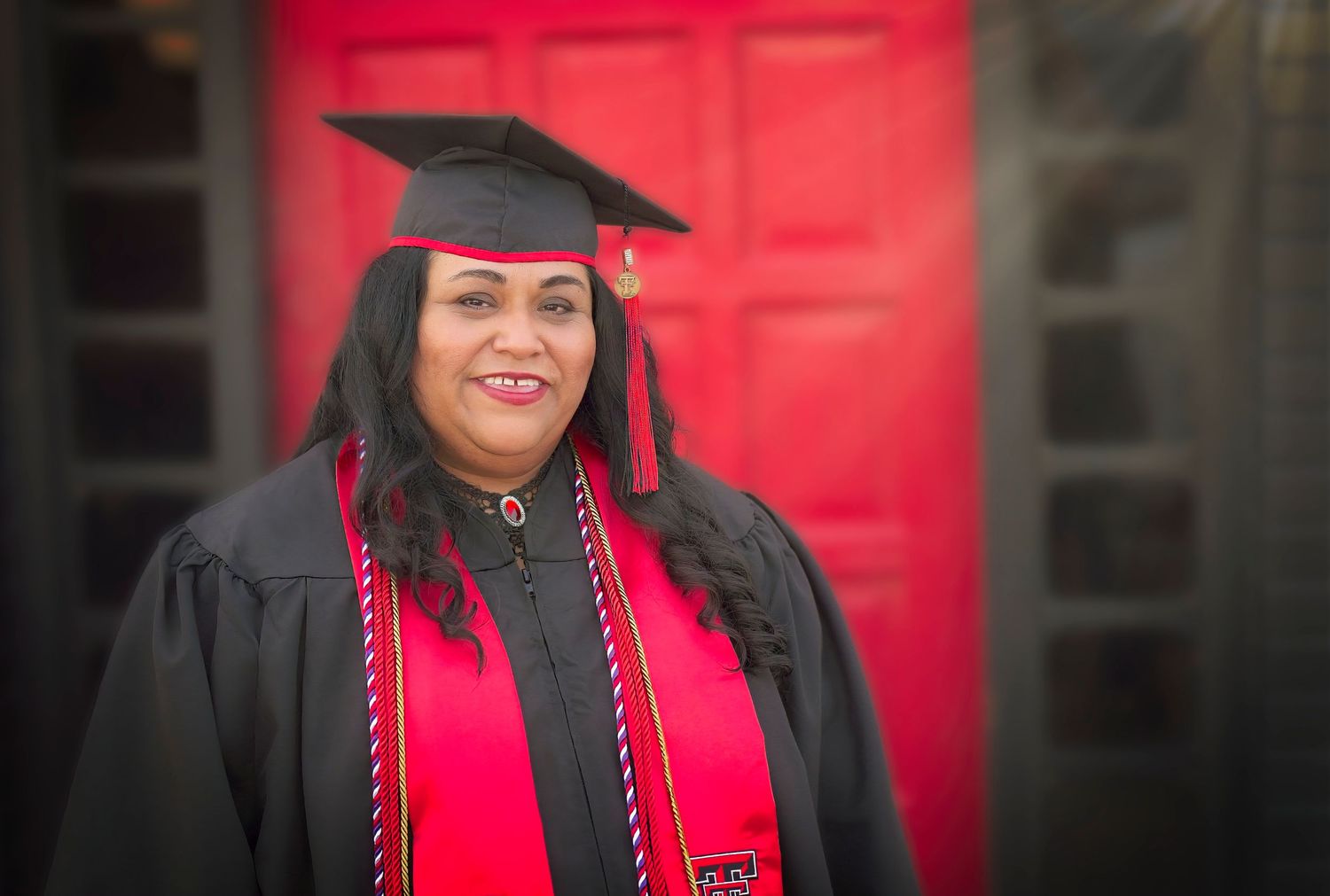 Graduate wearing red and black commencement regalia with decorative cord displays graduation cap against red backdrop.