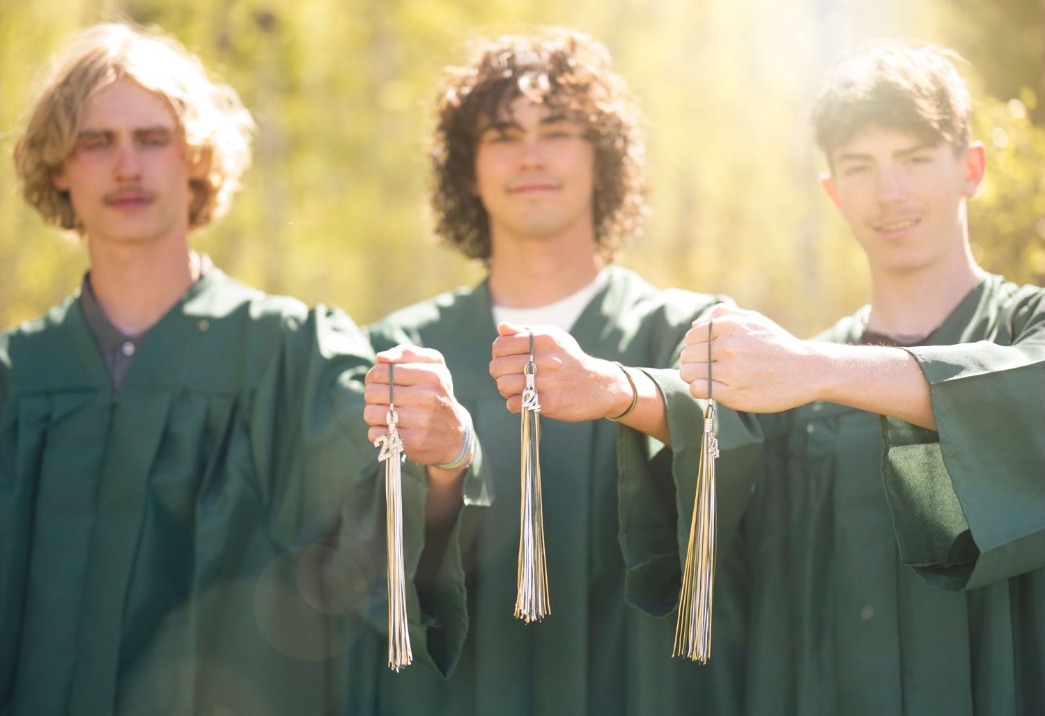 Row of graduates in green commencement robes standing together outdoors in bright sunlight.