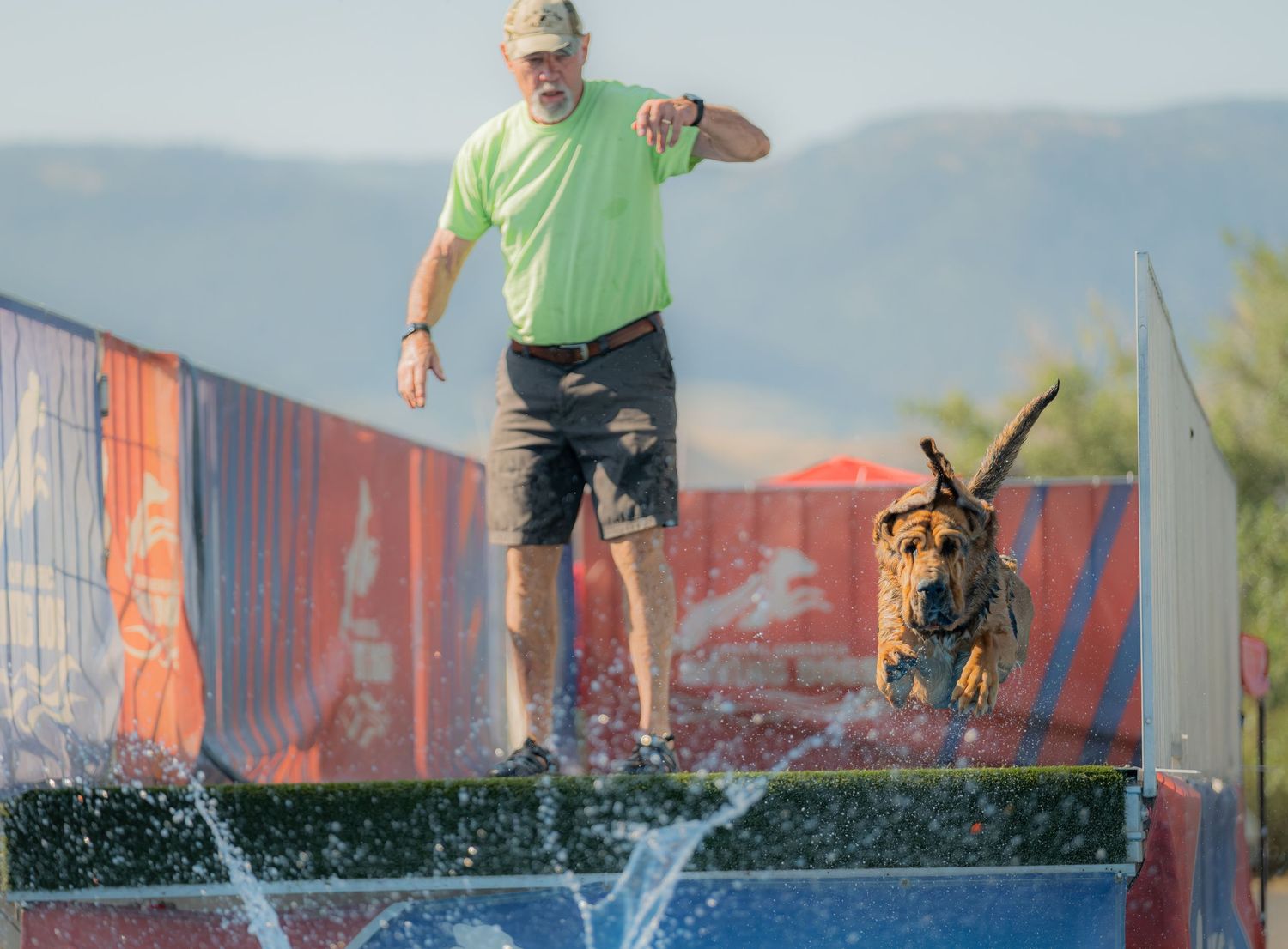 Man in green shirt with dog leaping into water at an outdoor dock diving competition.