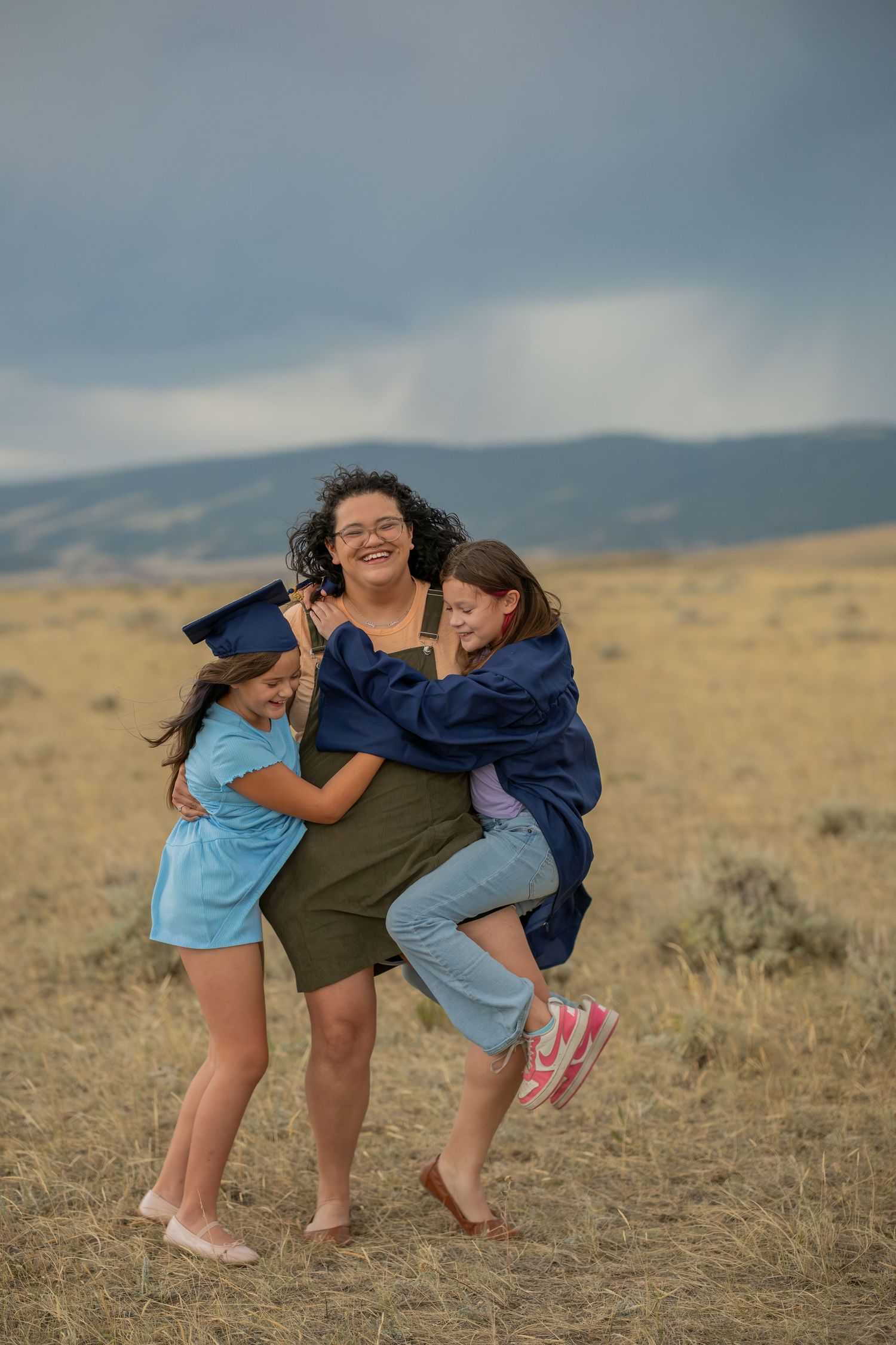 A woman in graduation cap and gown laughs joyfully as two young girls hug her in a golden field.