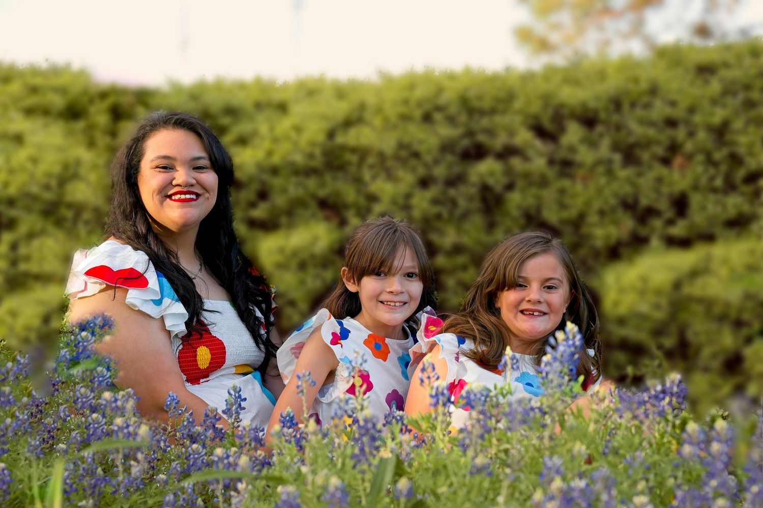 Three people in matching floral dresses smile while sitting among purple wildflowers.