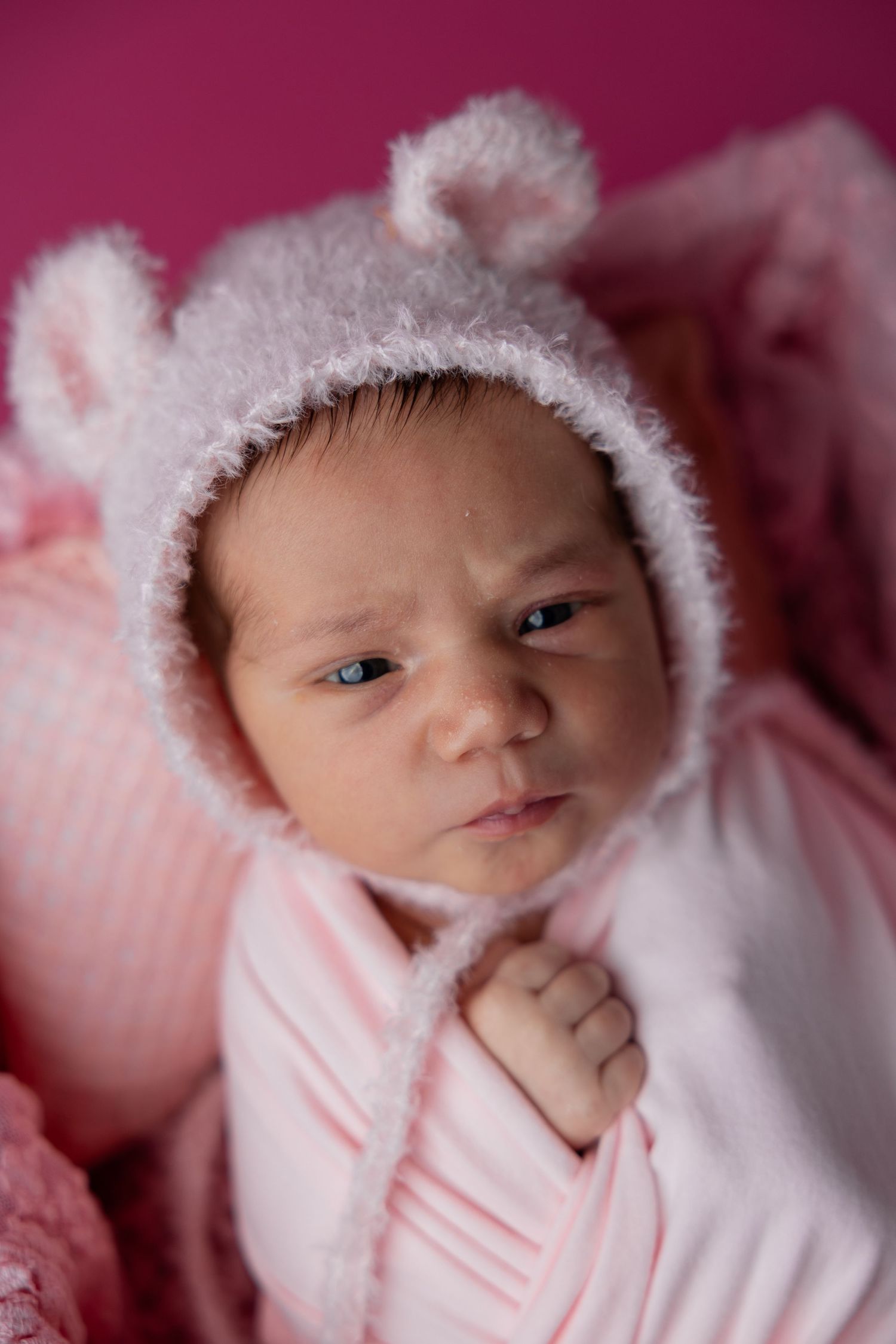Close-up of newborn baby in fuzzy bear bonnet wrapped in pink on pink background.