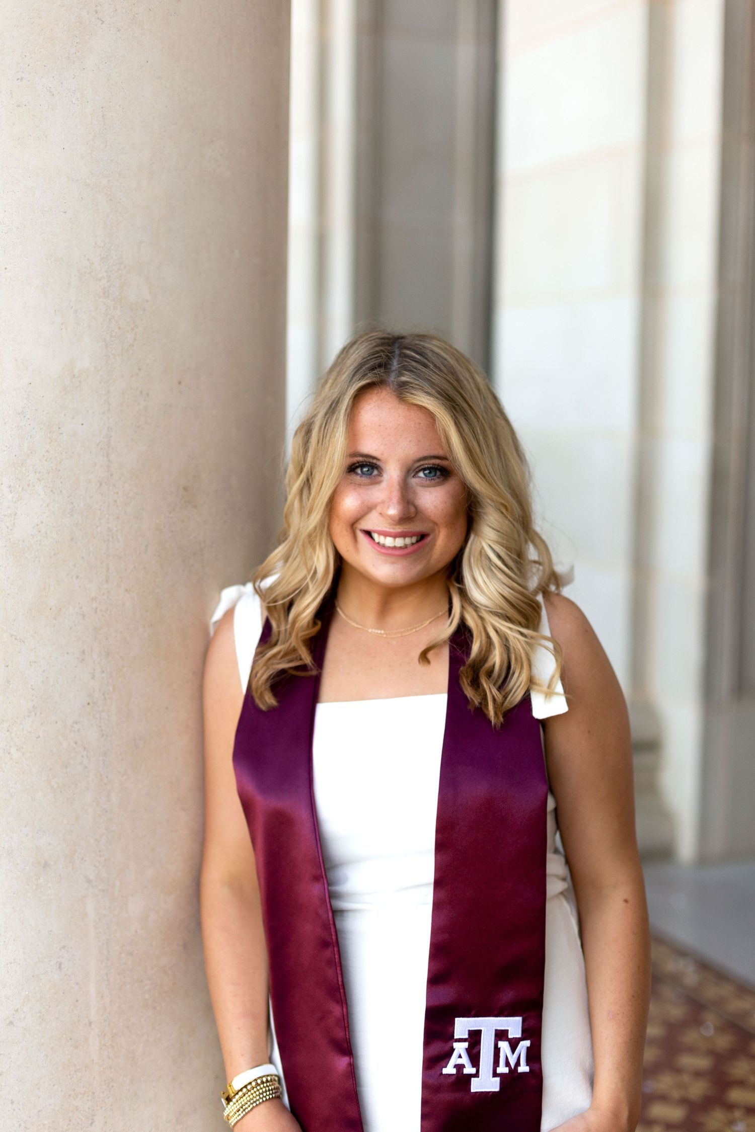 A graduate wearing a maroon Texas A&M University graduation stole over a white dress poses near a column.