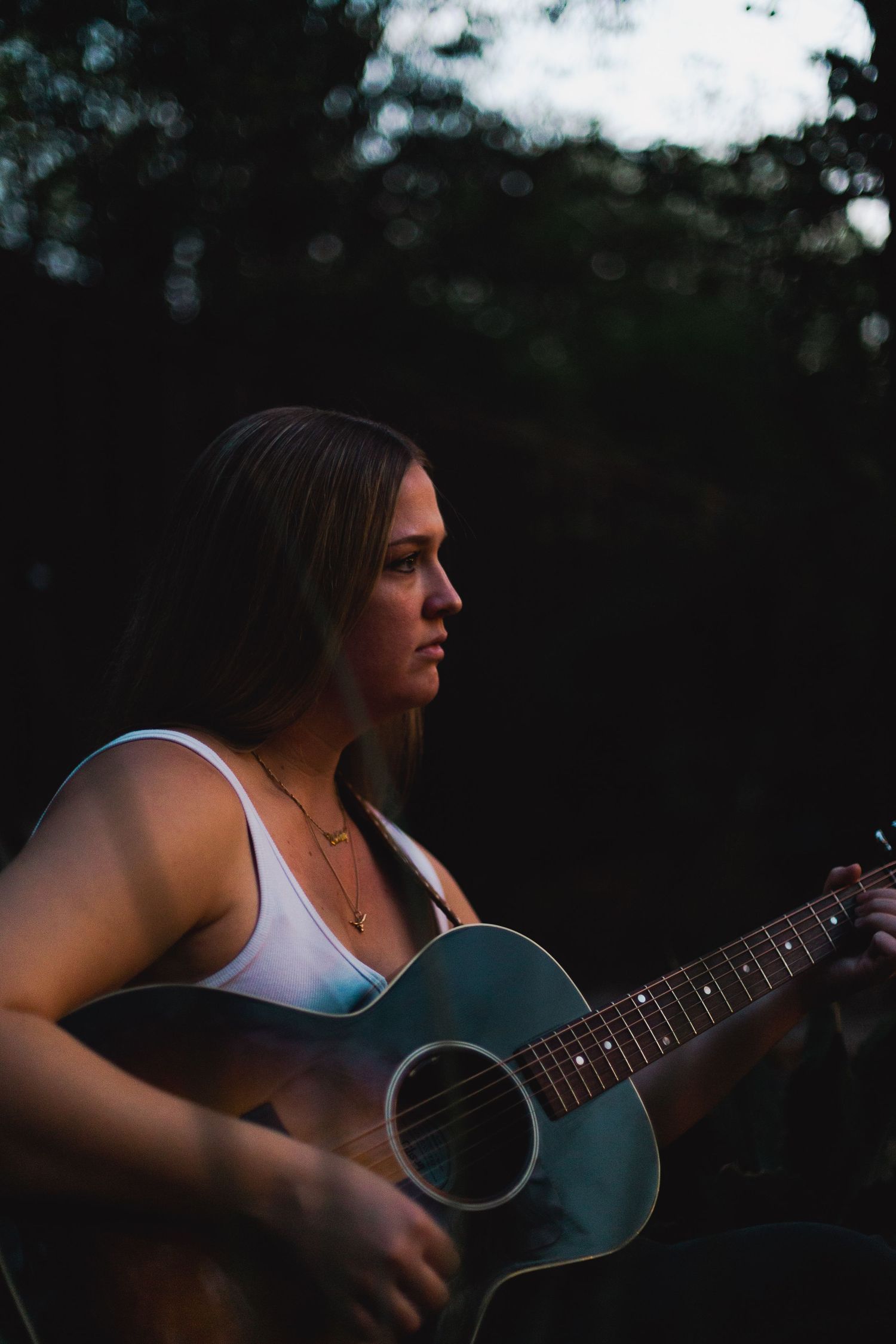 A musician playing acoustic guitar outdoors at dusk while wearing a white tank top.
