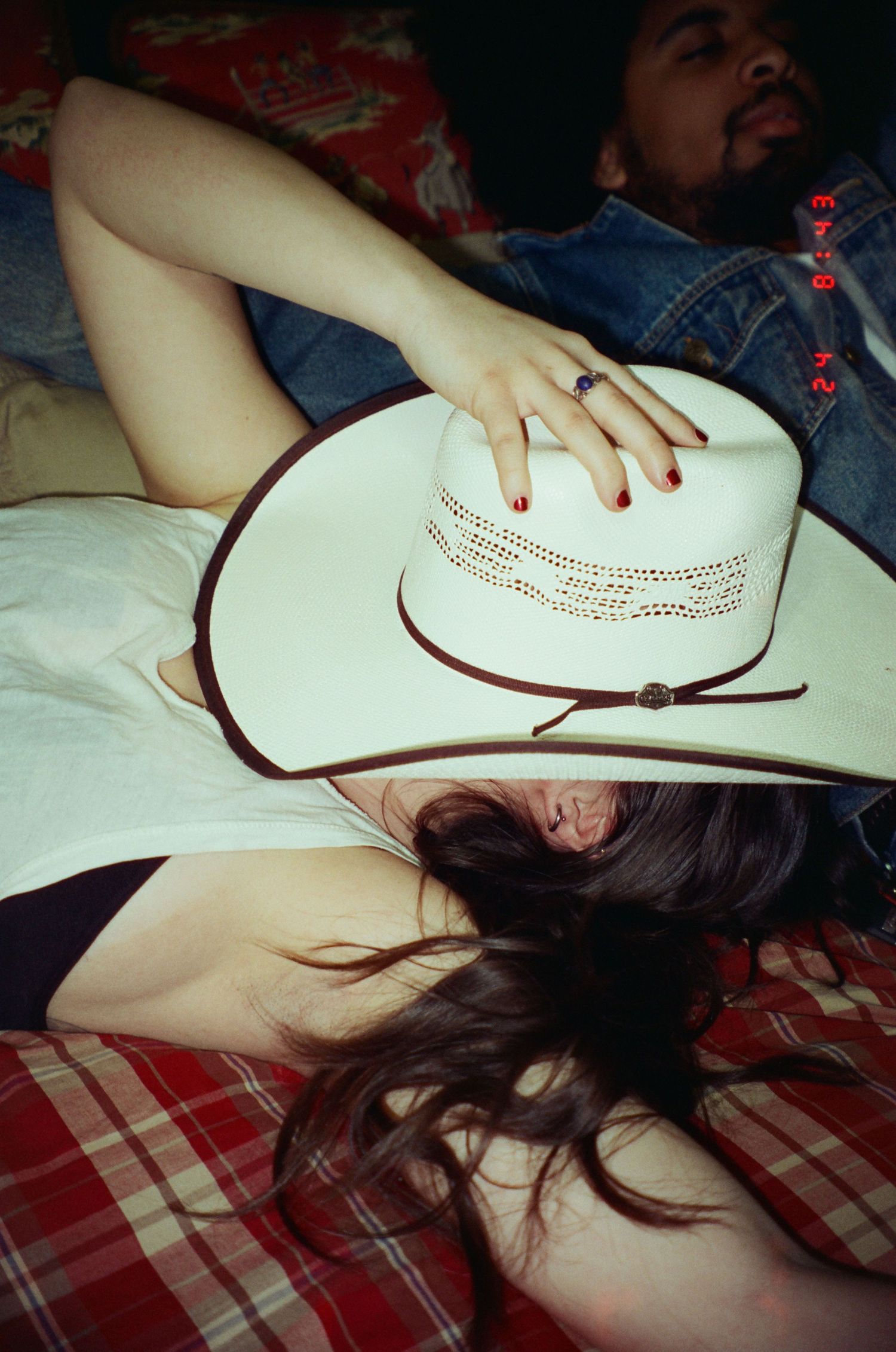 A white cowboy hat rests on someone lying on a plaid patterned bed with dark hair spread out.