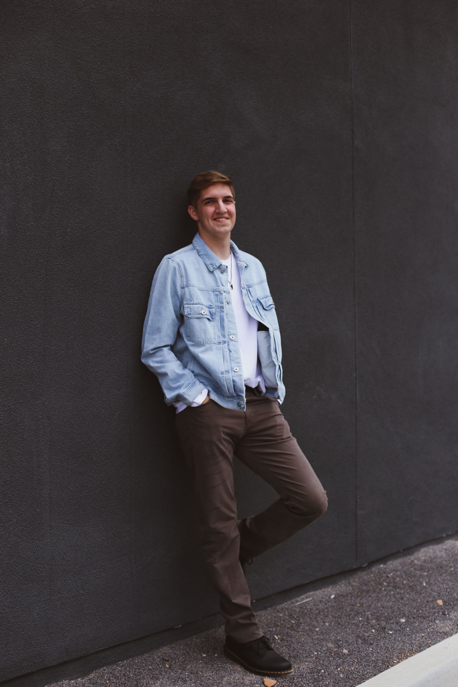 A man in light denim jacket and brown pants poses casually against a dark gray wall outdoors.