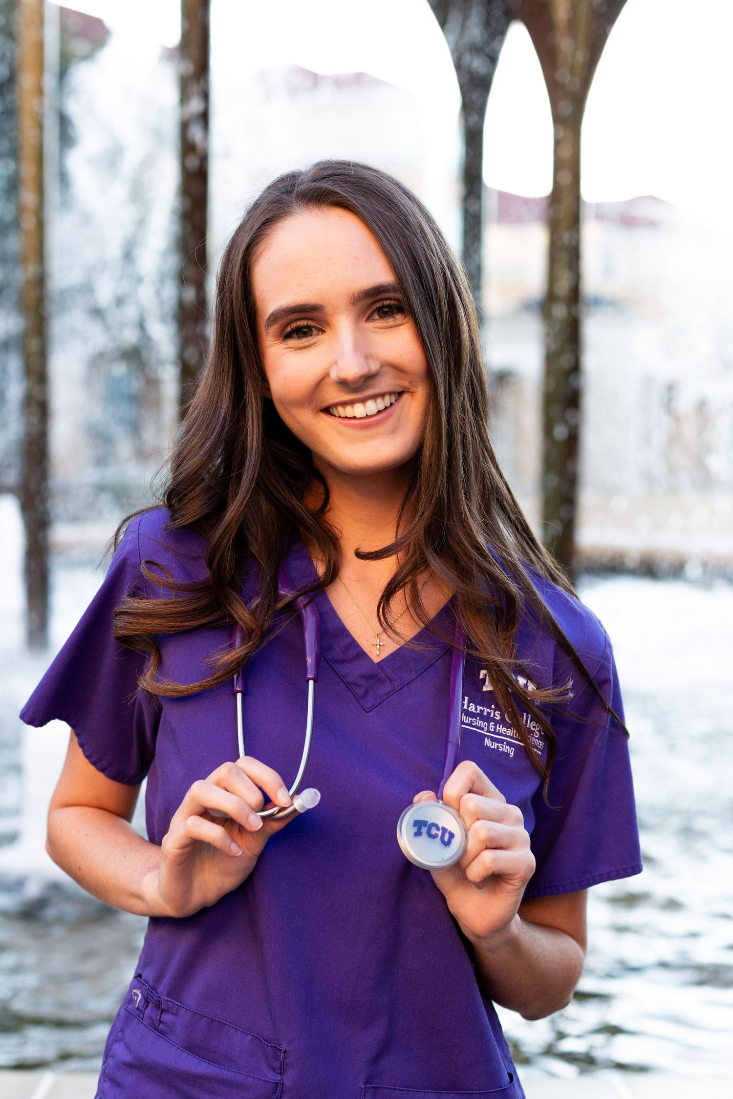 A healthcare professional in purple scrubs holding medical equipment stands outdoors with a bright smile.