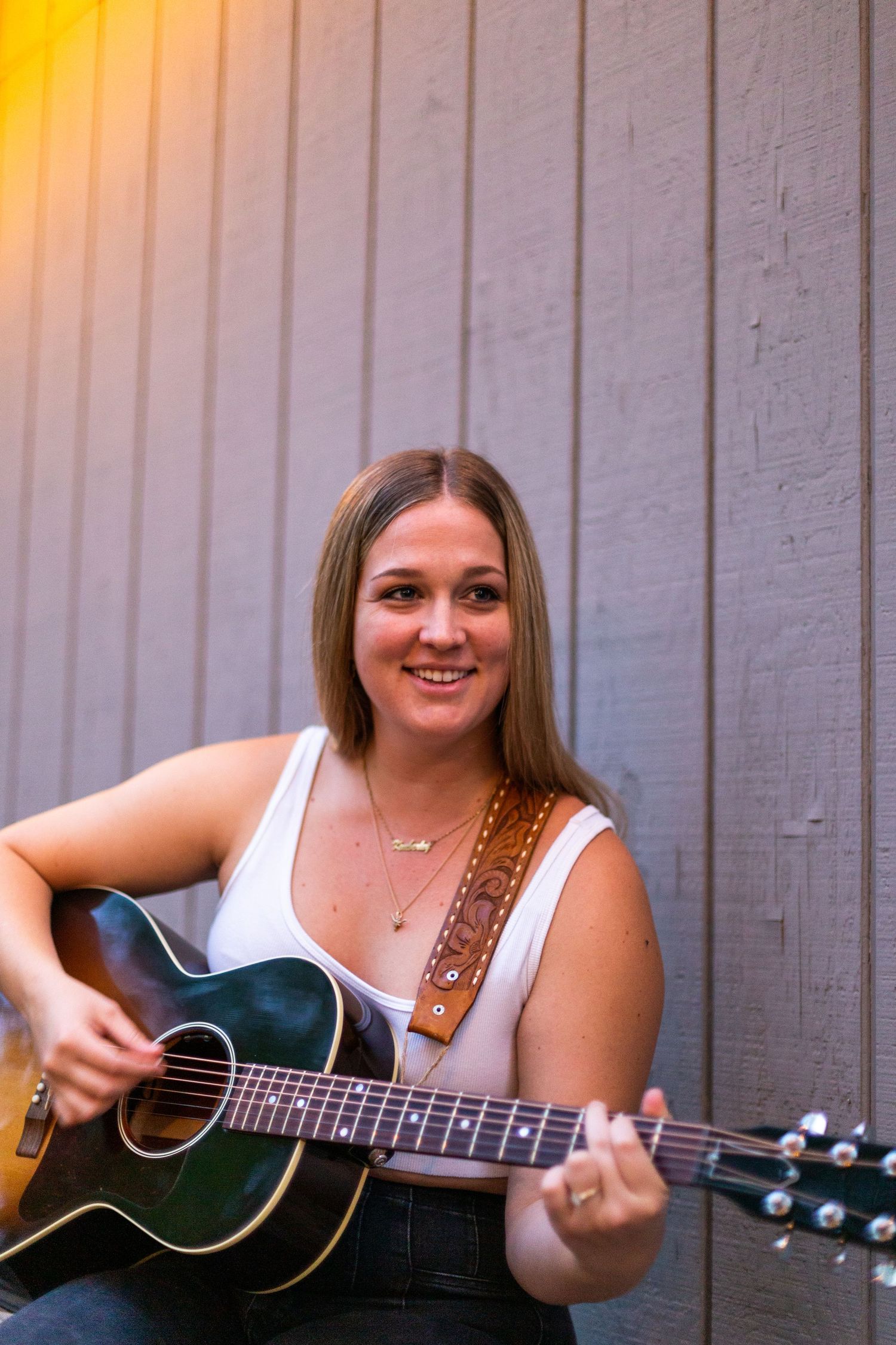 A musician smiling while playing acoustic guitar against a grey wall with warm lighting.