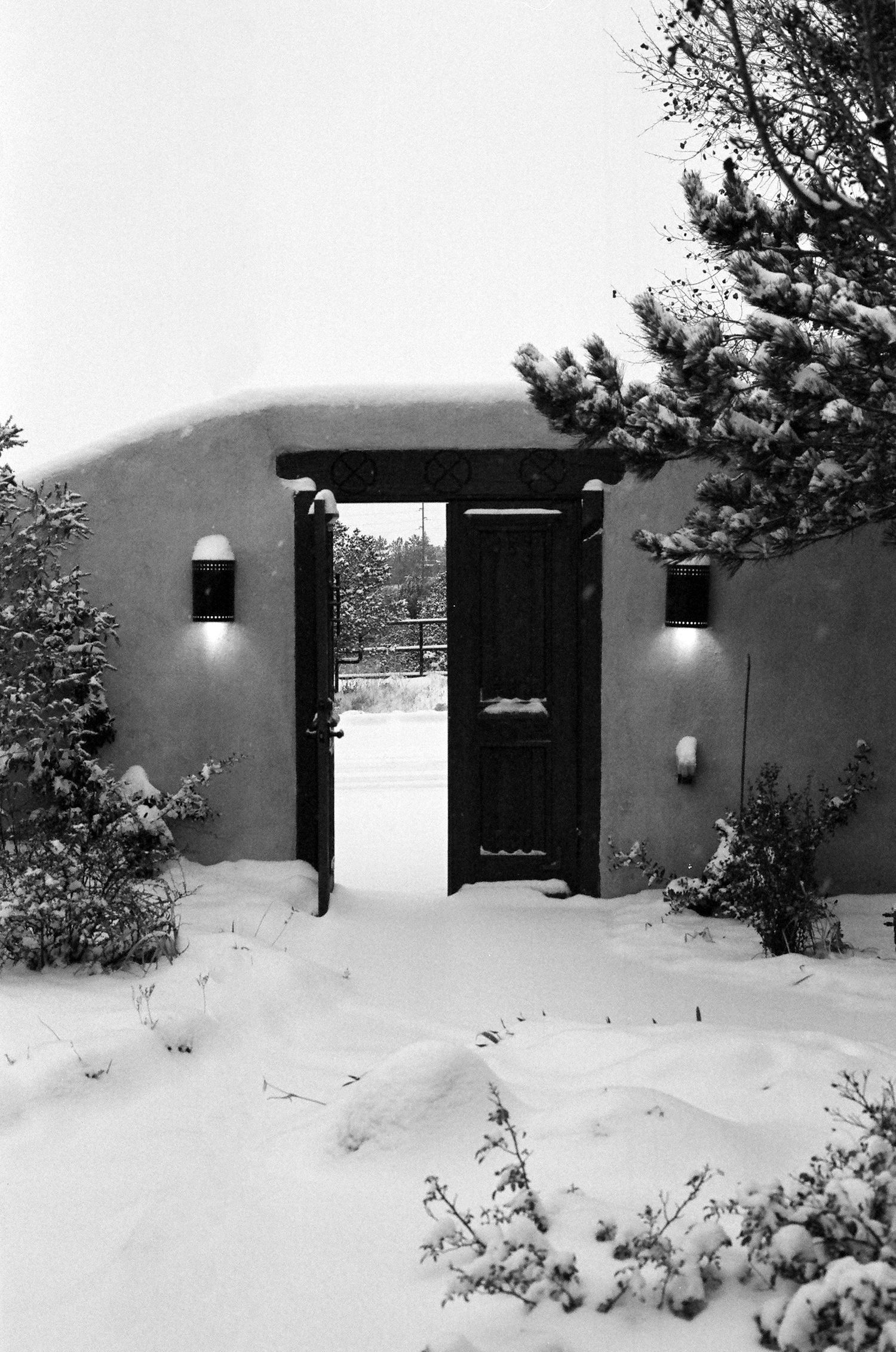 A snowy entrance to an adobe-style home with wall lights flanking a dark wooden door.