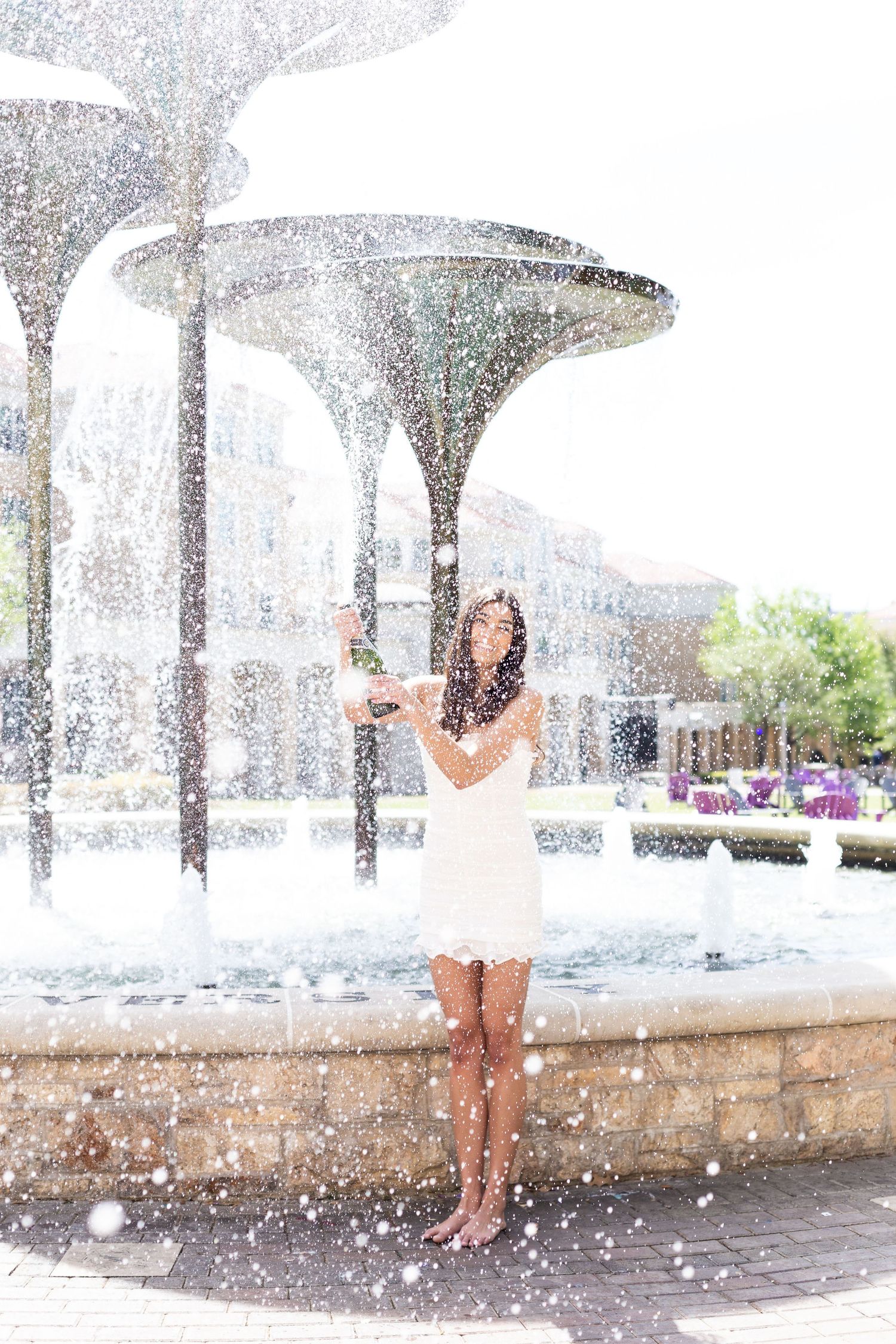 A person in a white dress stands under outdoor water features spraying mist on a summer day.