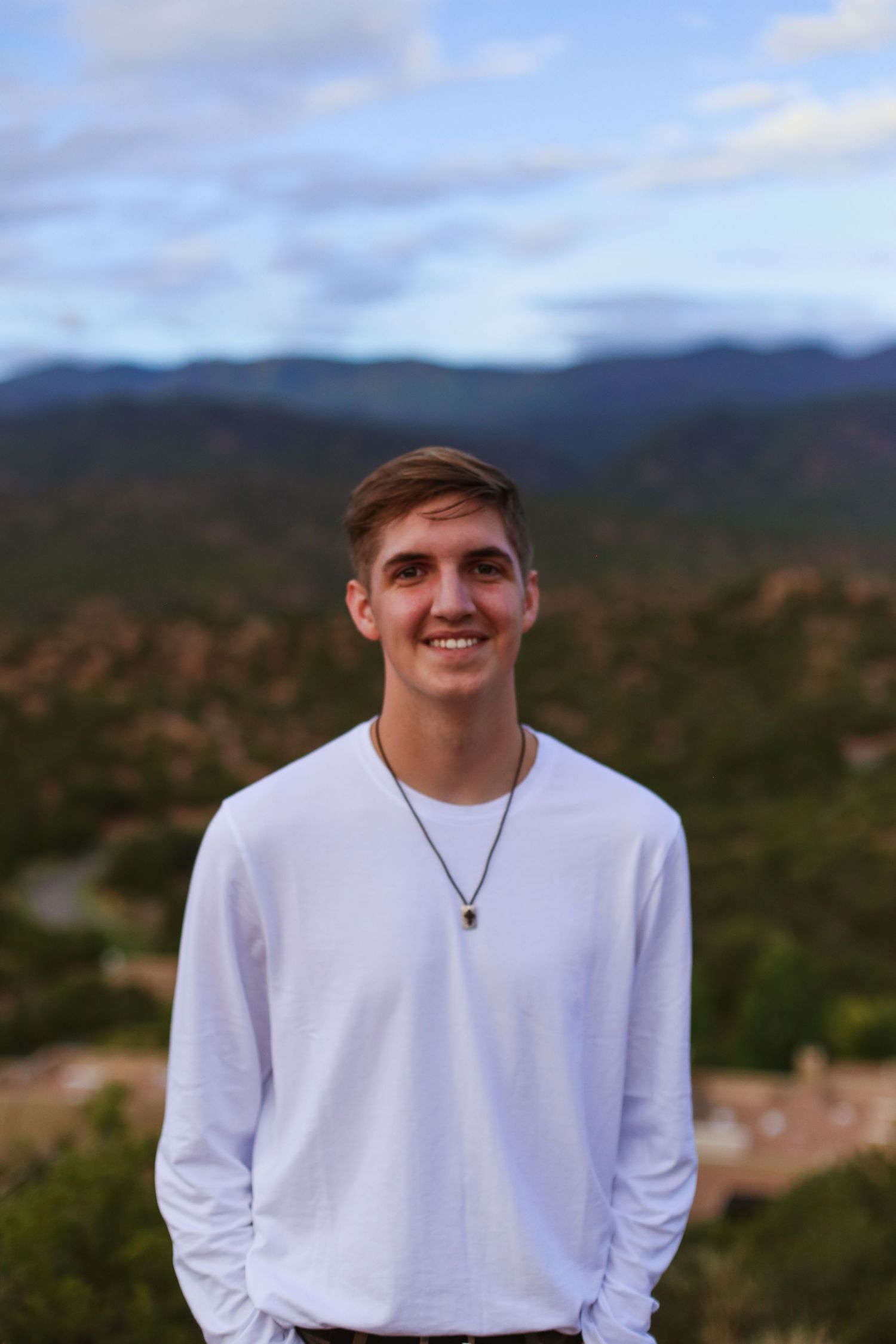Young person in white long sleeve shirt and necklace poses outdoors with mountains and cloudy sky in background.