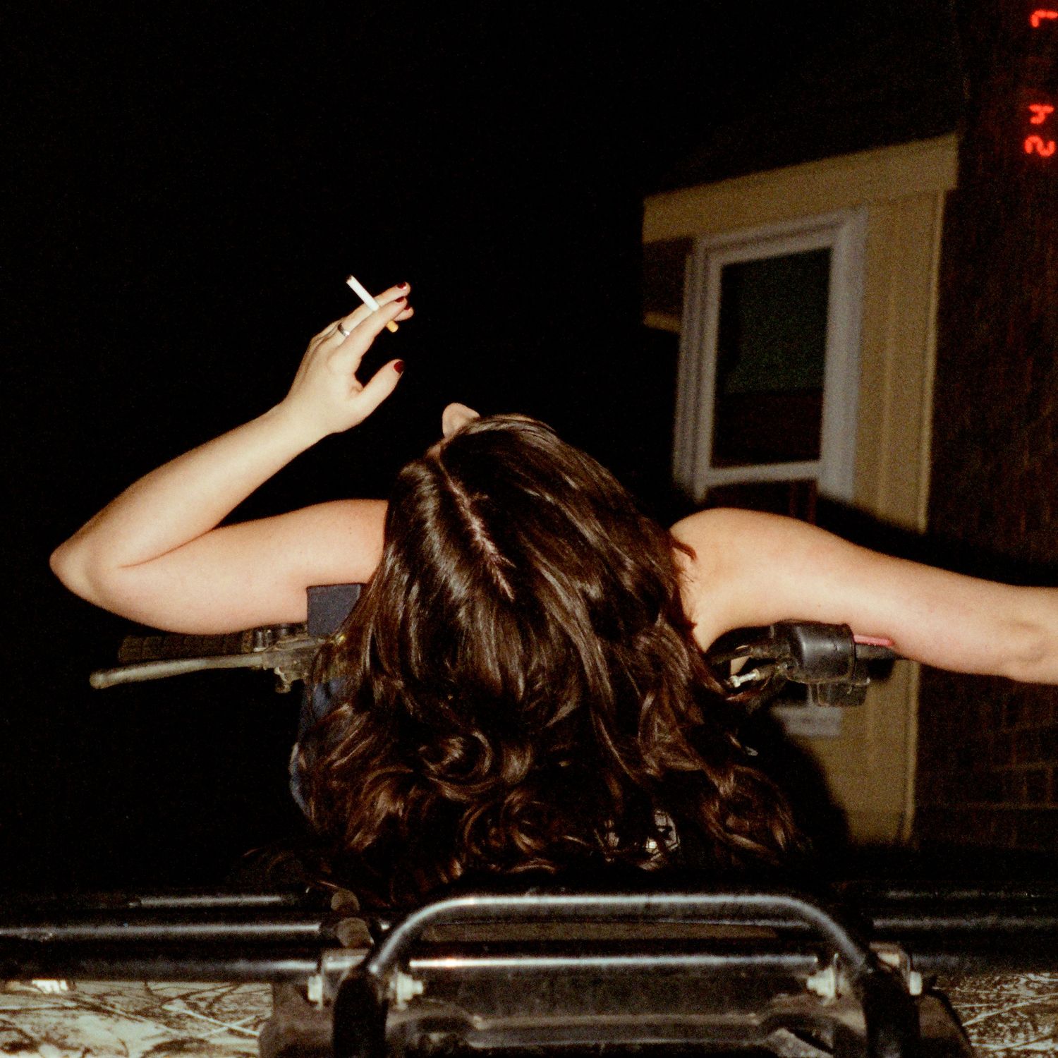 Back view of person with dark wavy hair raising their arms against a dark night sky while leaning on a railing.
