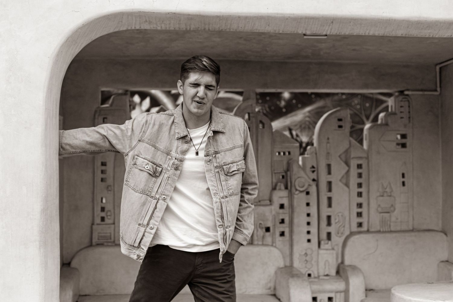 A person in a denim jacket poses against a curved architectural backdrop.