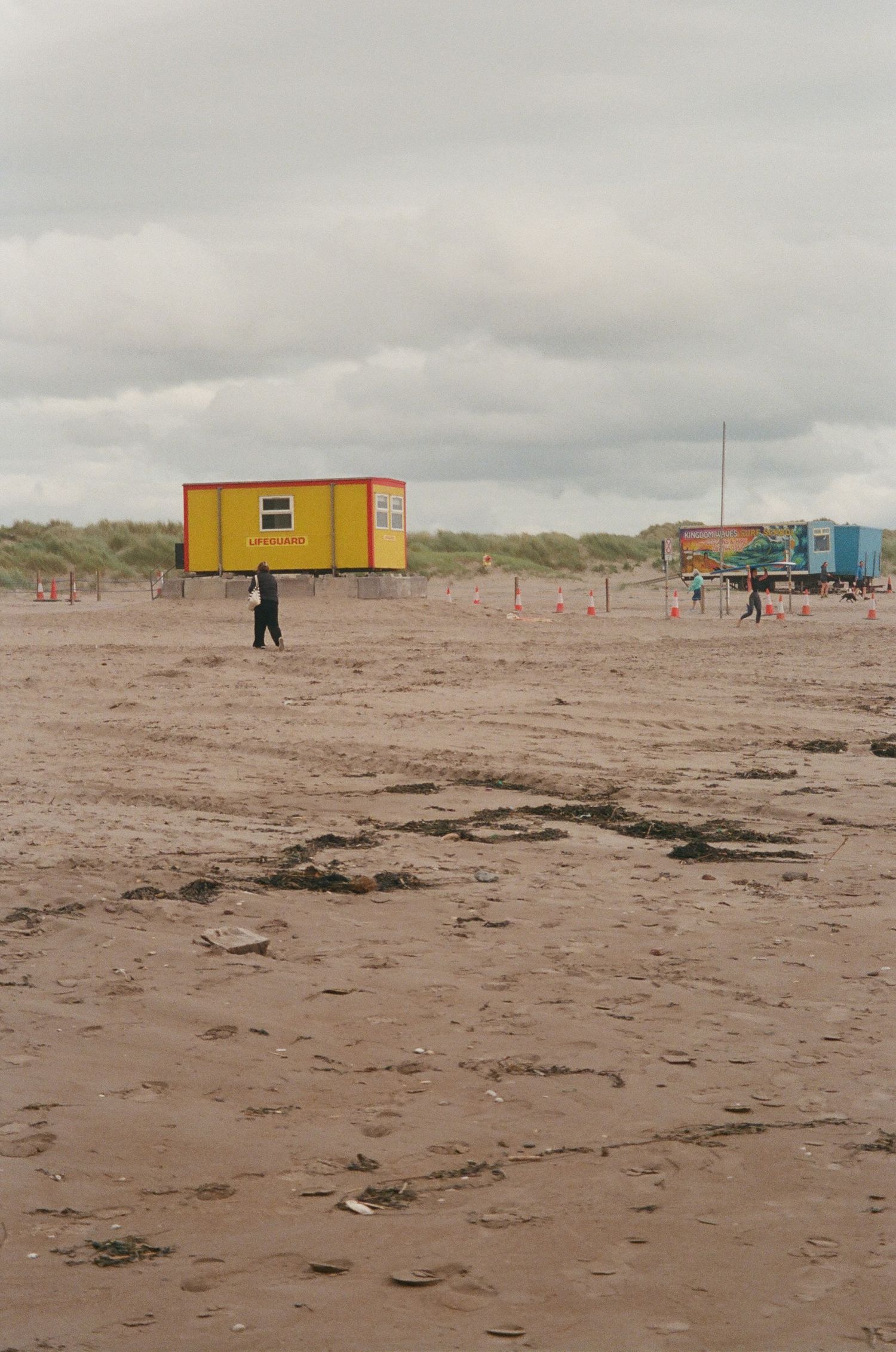 Small yellow and blue beach huts stand on a desolate sandy shore under overcast skies.
