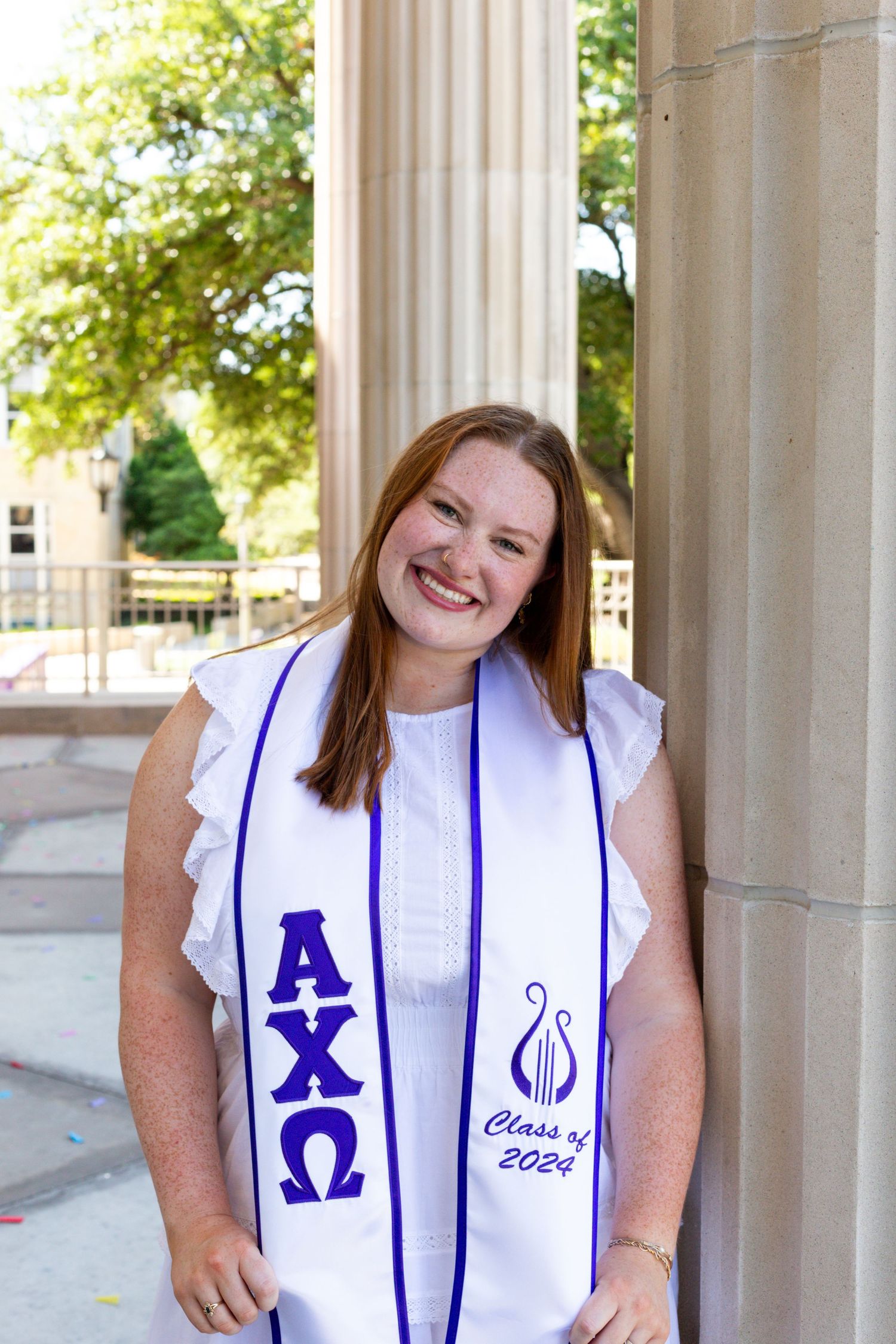 A graduate wearing white graduation stole with blue Alpha Chi Omega letters and lyre symbol stands smiling by a column.