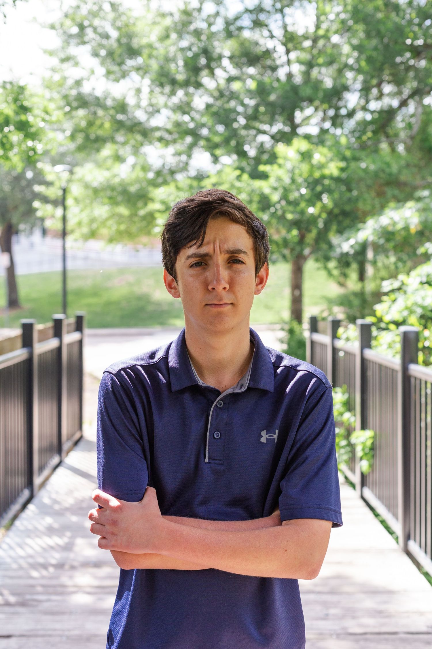 A person in a navy blue Under Armour polo stands with crossed arms on a wooden walkway surrounded by greenery.