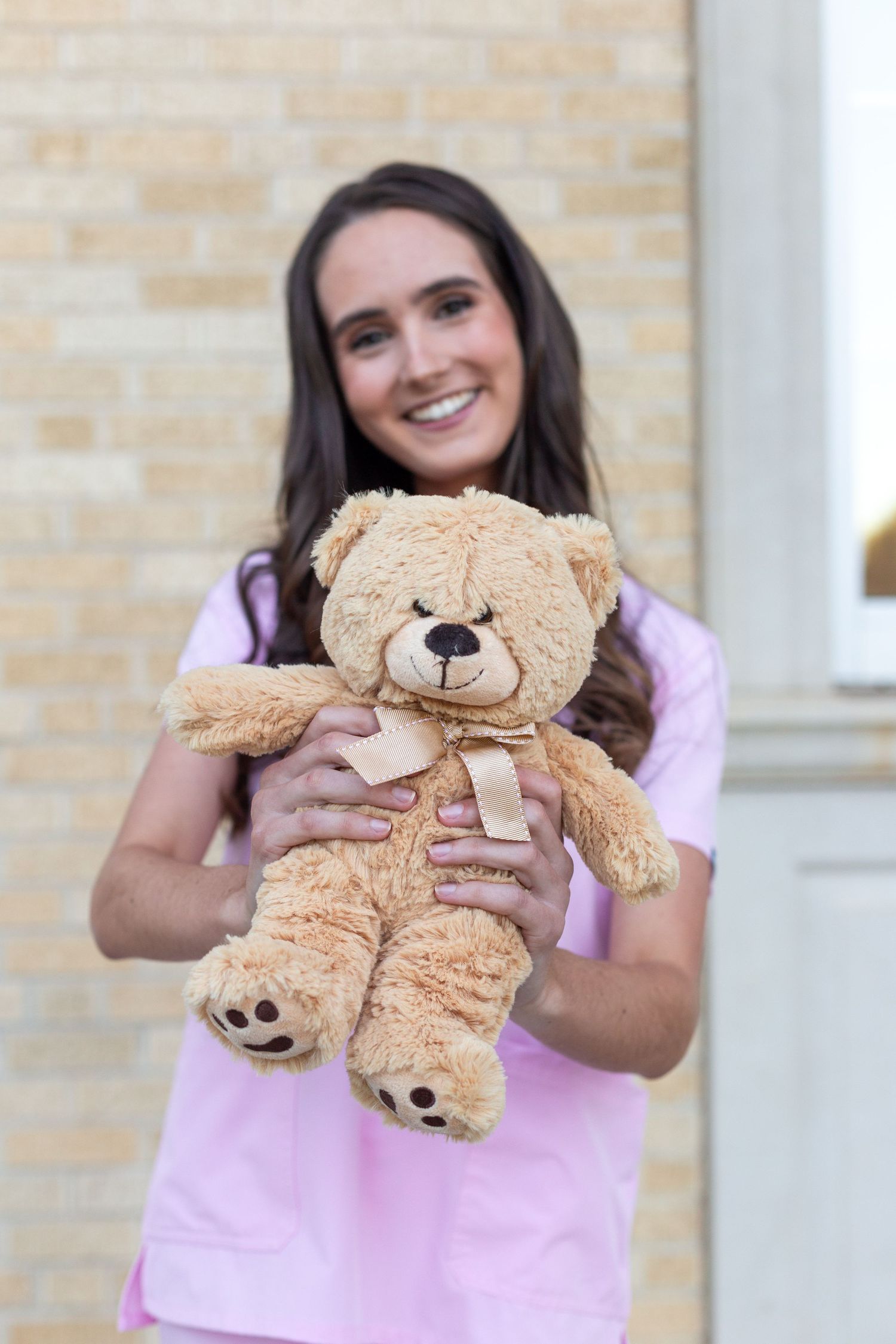 Someone in a pink t-shirt holds a cuddly tan teddy bear with a bow while standing in front of a brick wall.
