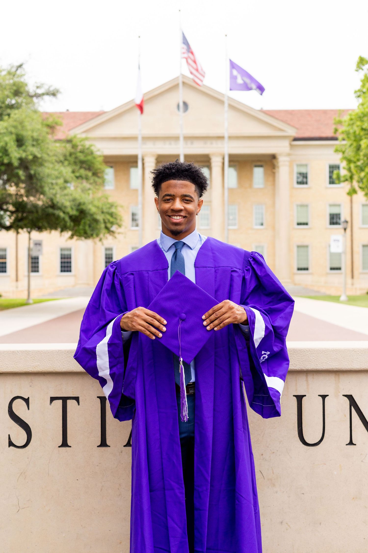 Graduate in purple academic regalia stands proudly outside university building with American flag in background.