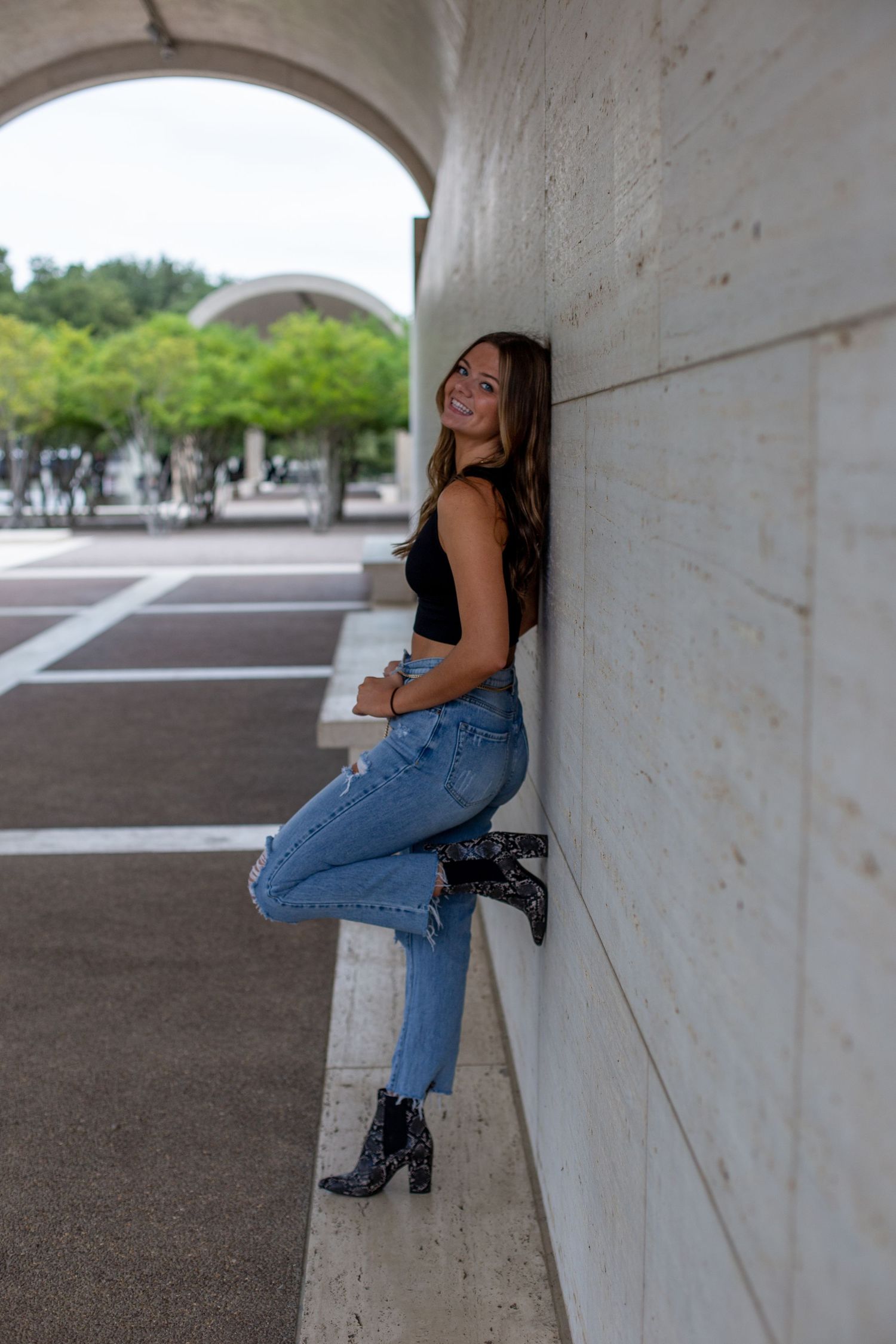 A person in blue jeans and black boots poses against a white concrete wall under architectural arches.