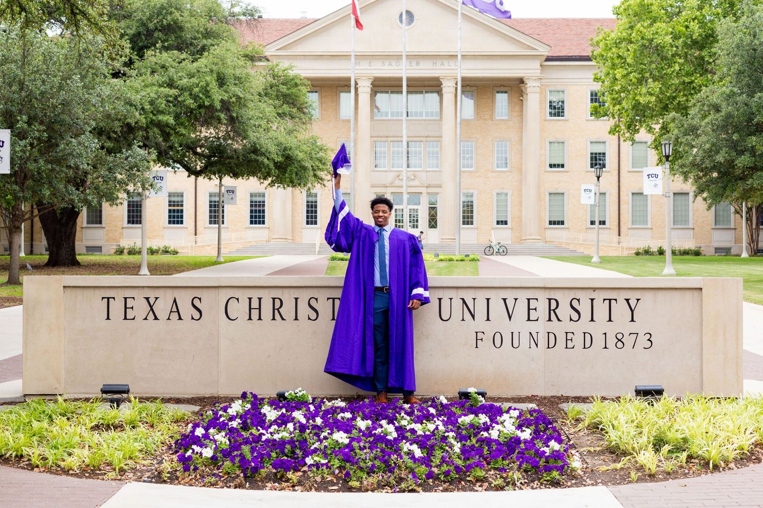 Graduate in purple regalia poses in front of Texas Christian University welcome sign with purple flowers in bloom.