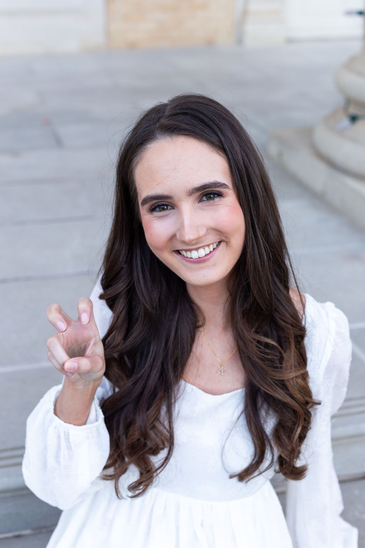 A graduate in a white blouse makes a peace sign while smiling at the camera.