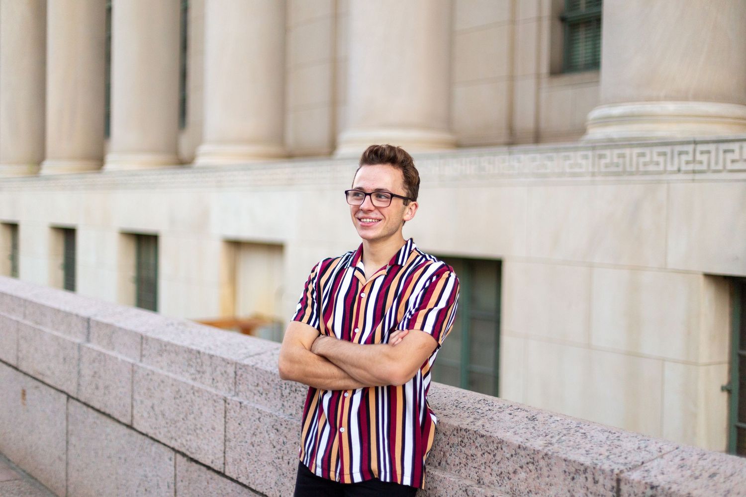 Someone in a striped shirt smiles while leaning against a stone wall with Greek columns in the background.