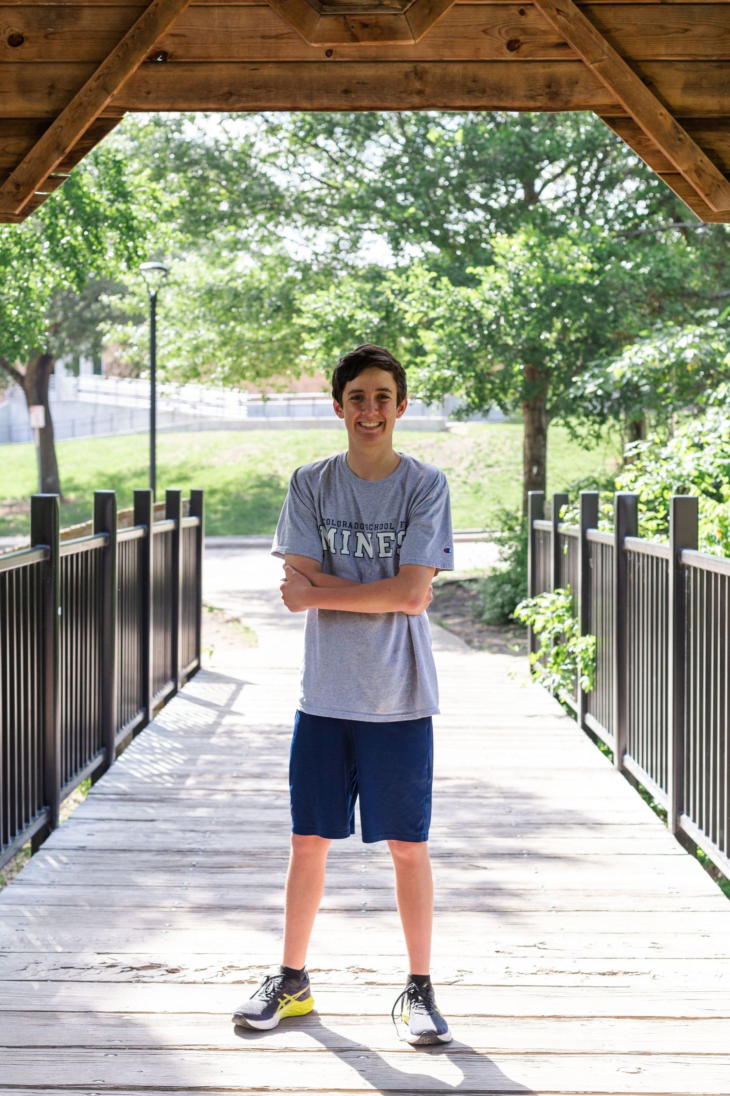 Someone in a UNC t-shirt and navy shorts stands smiling on a wooden bridge walkway surrounded by greenery.