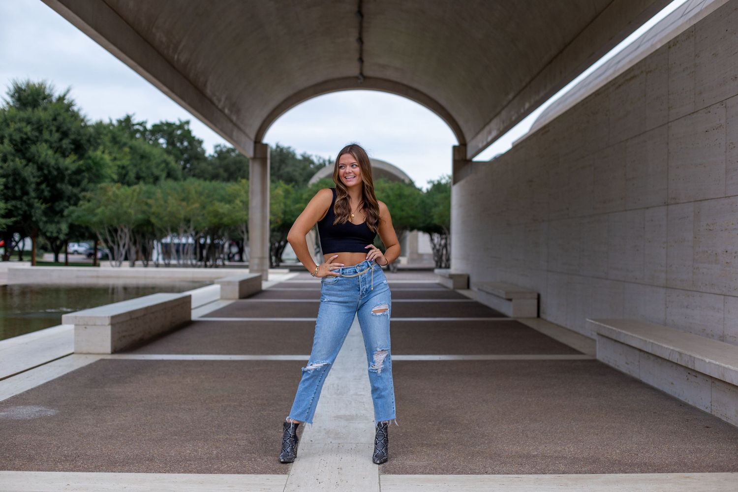 A person in casual attire poses confidently under a concrete archway on a sunny day.