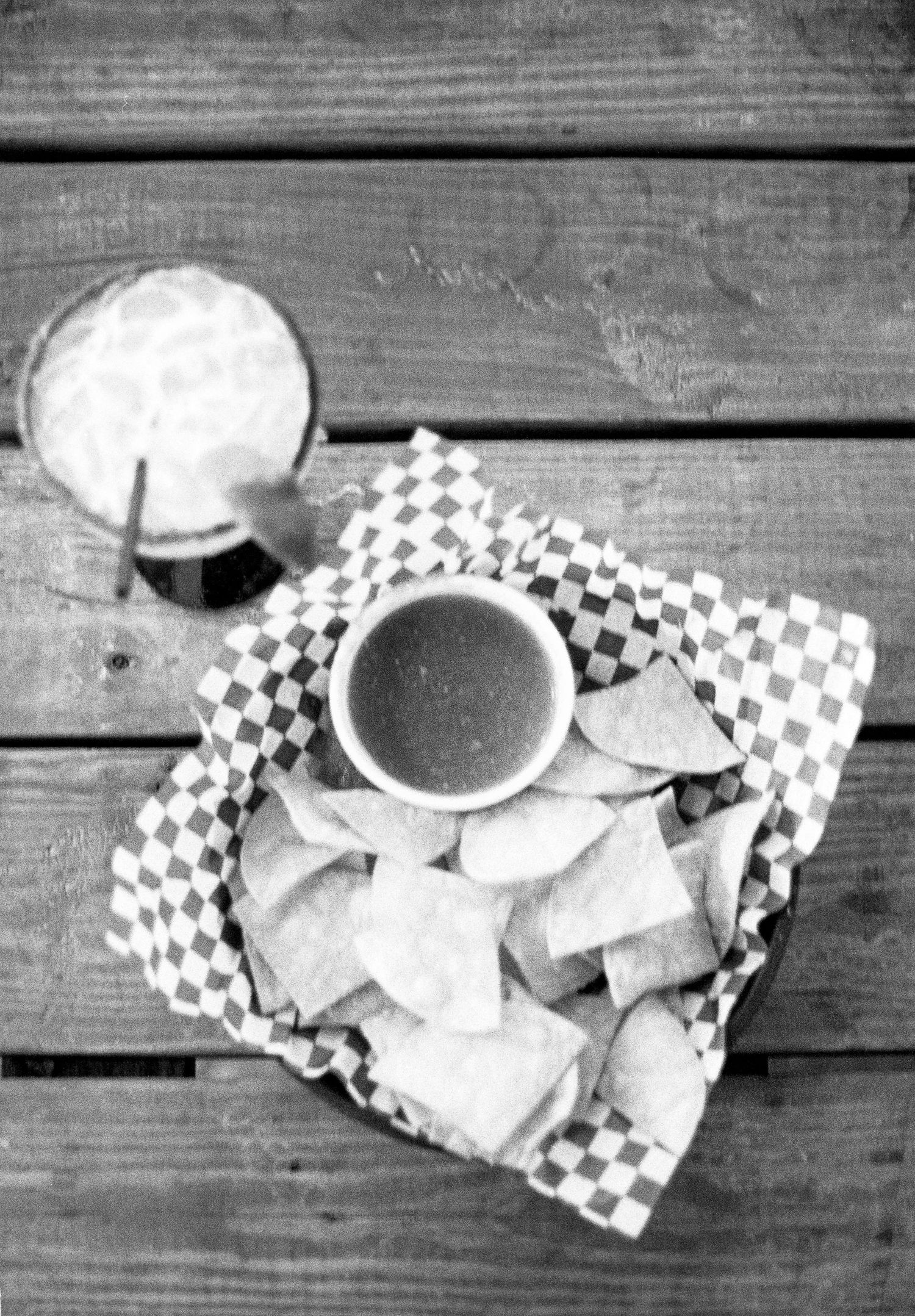 Black and white photo of tortilla chips and salsa served in a checkered paper lined basket with a drink on wooden table.