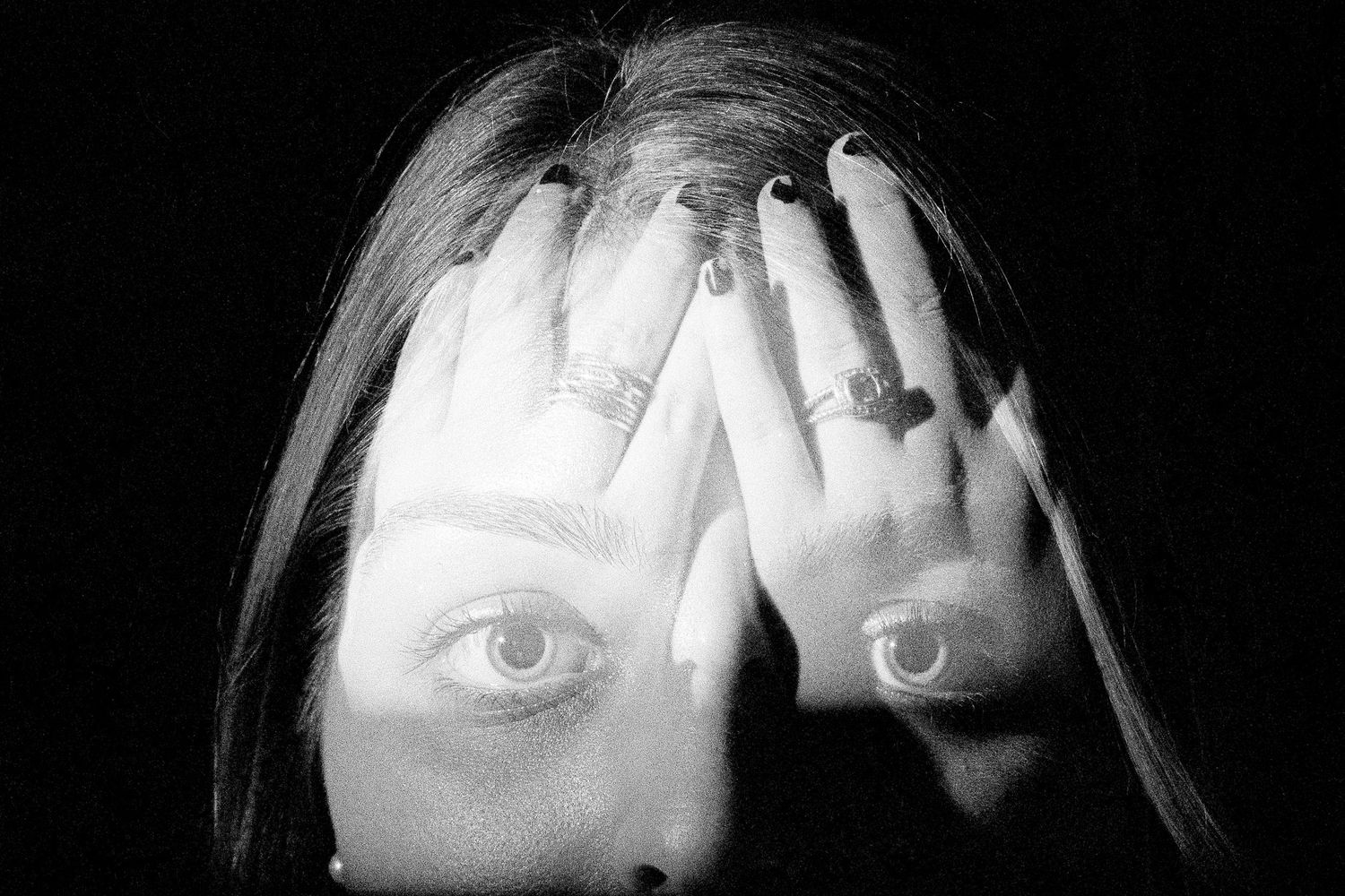 Black and white artistic photograph showing eyes painted on palms covering the face, creating surreal double vision effect.
