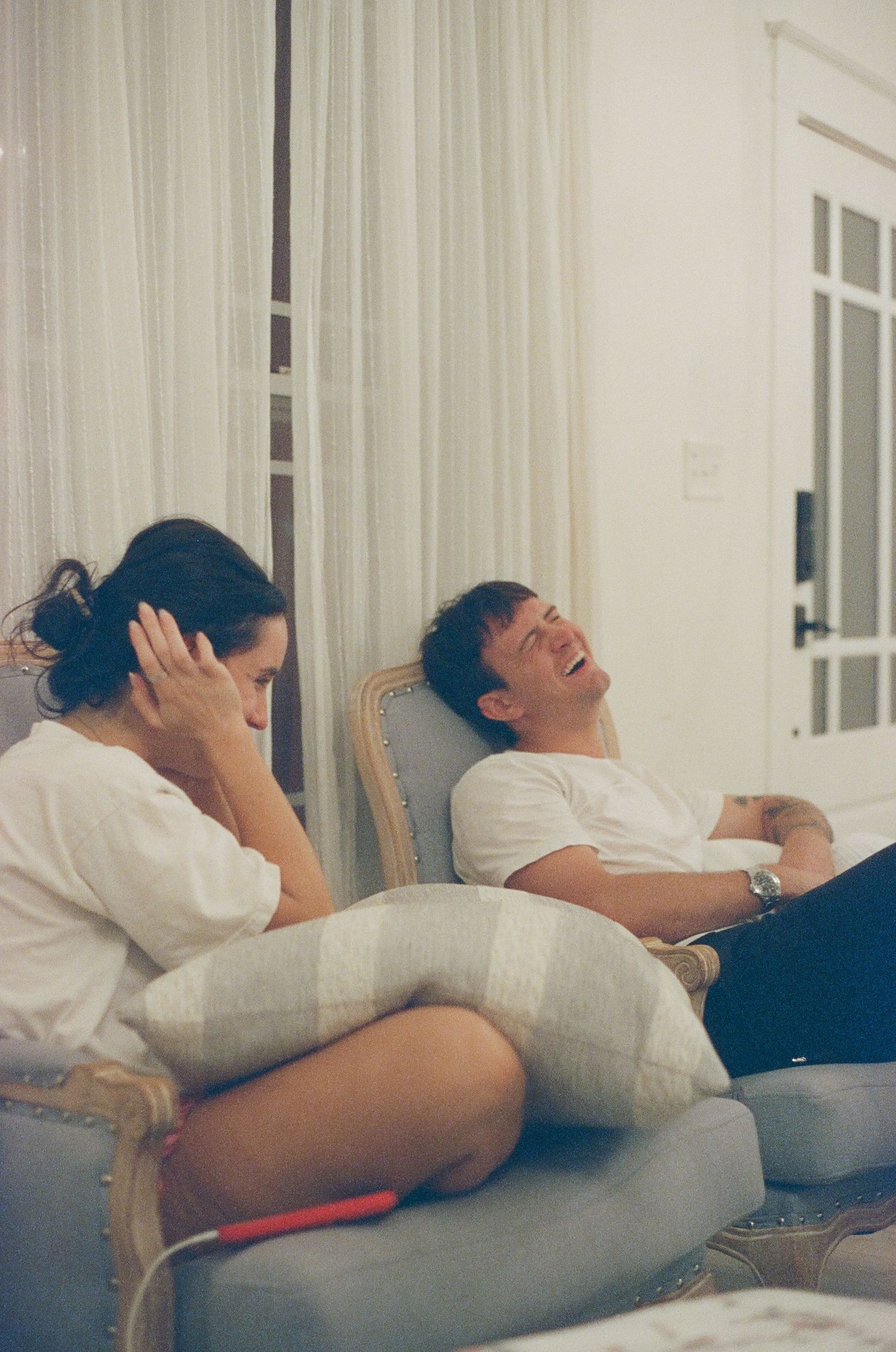 Two people relaxing on a couch, one appears frustrated while the other laughs heartily in a bright living room.