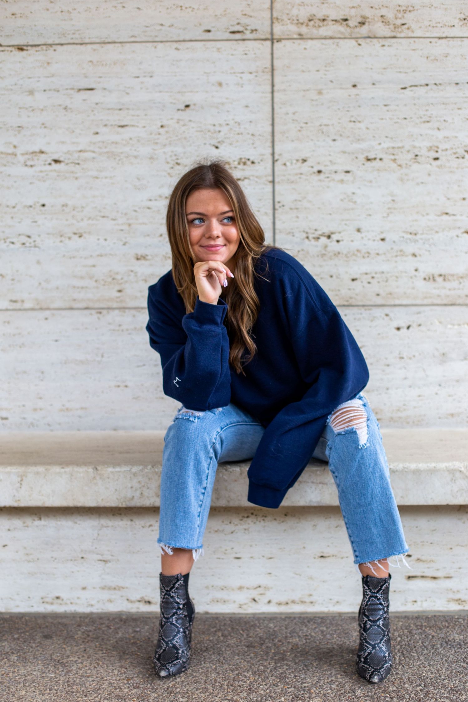 Someone wearing a navy sweater and distressed jeans sits casually on concrete steps with snakeskin boots.