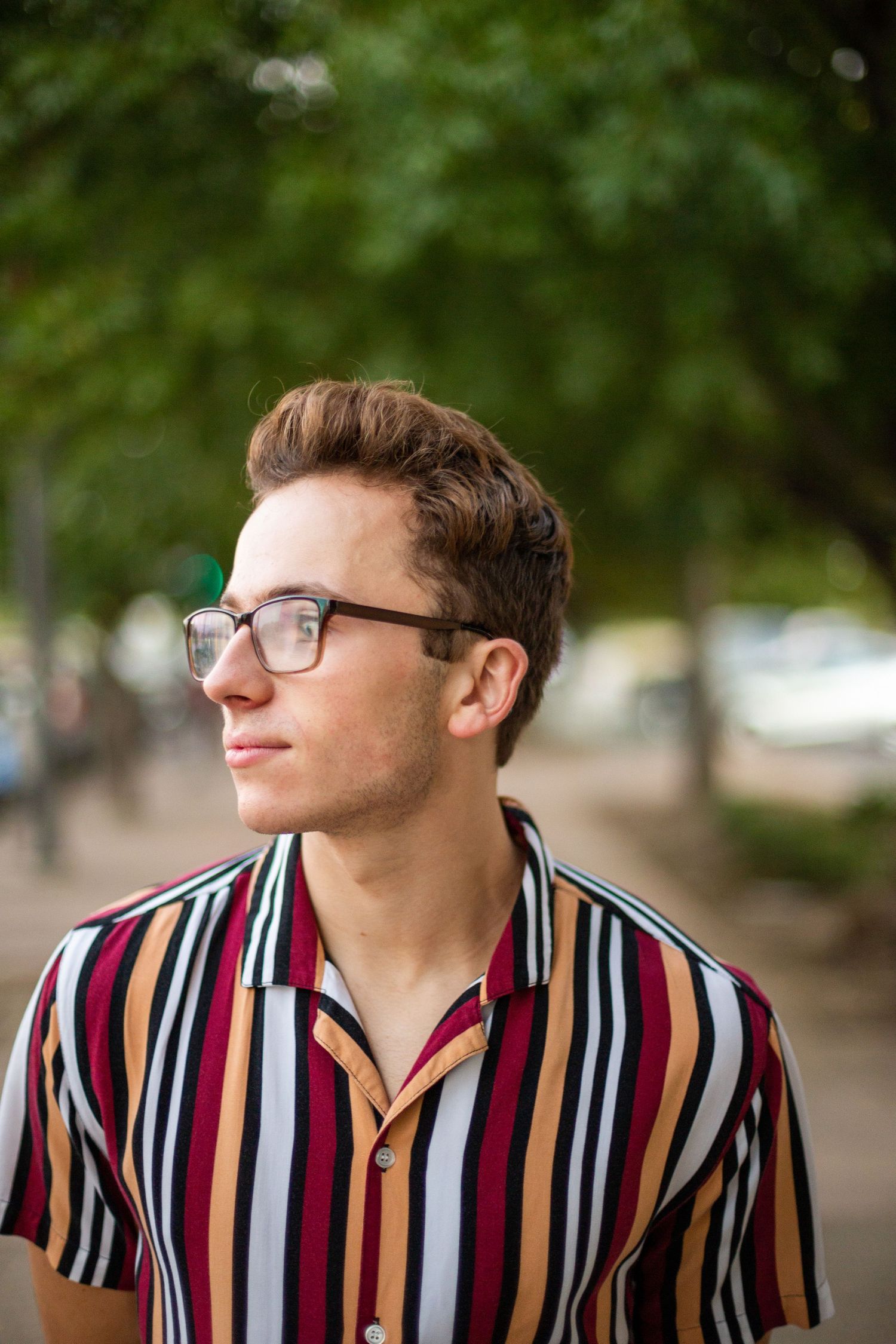 A person wearing glasses and a colorful striped short-sleeve button-up shirt looks off into the distance outdoors.