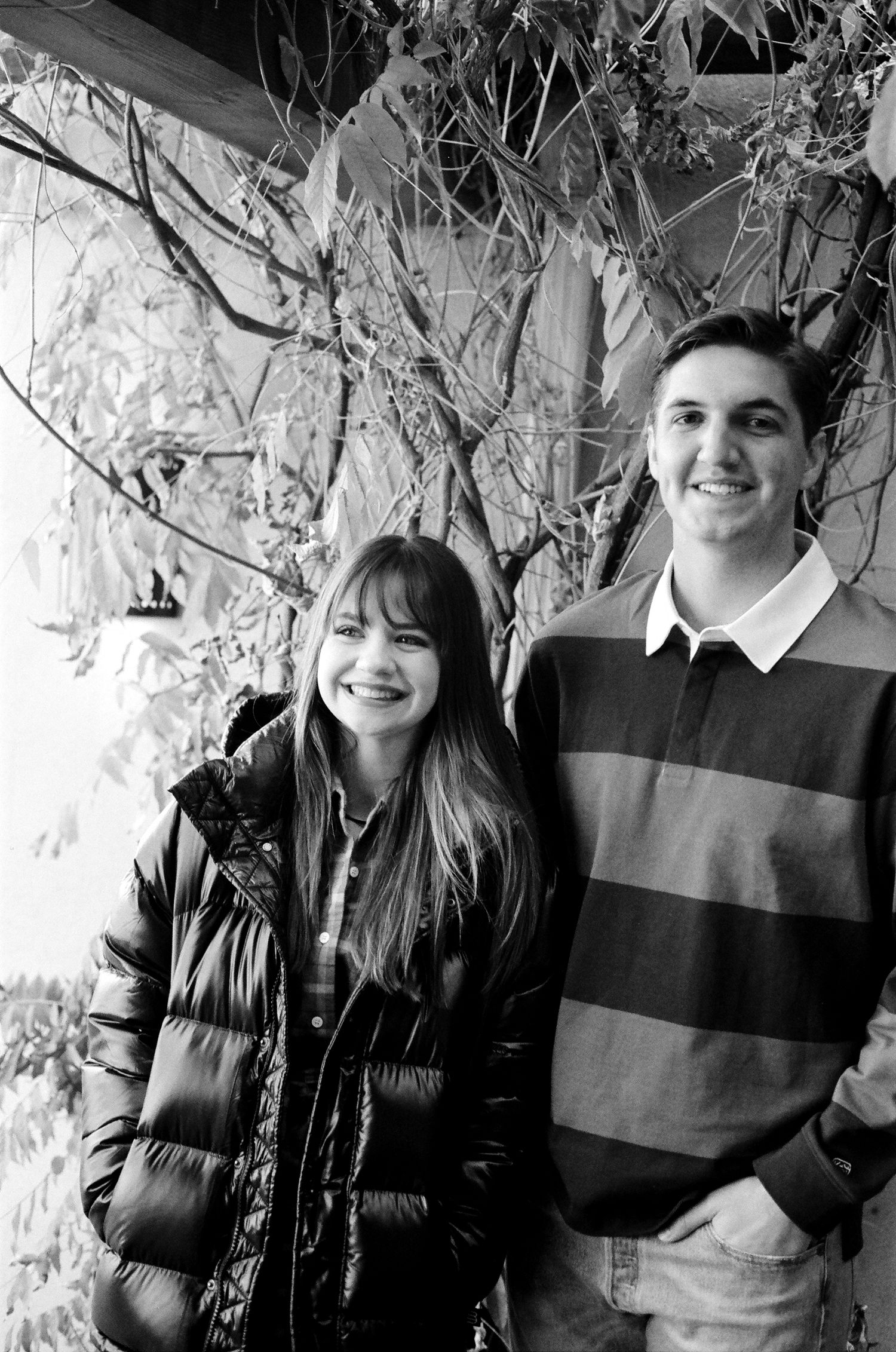 Black and white photo of a couple standing together outdoors with leafy vines visible in the background.