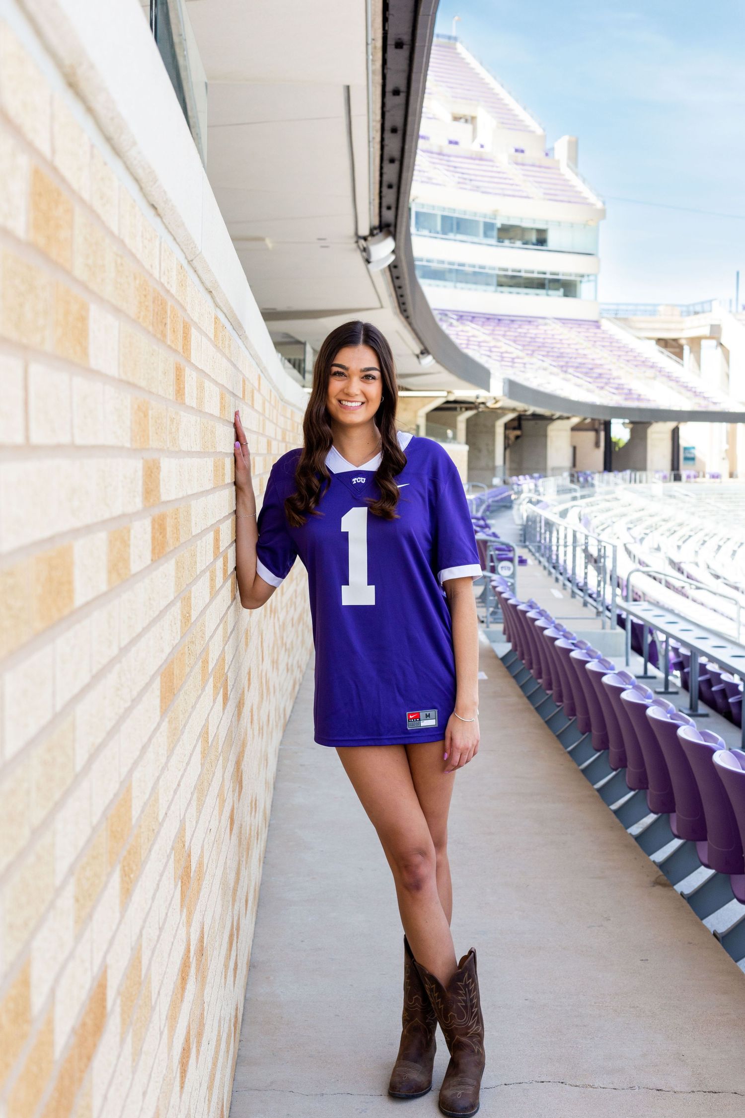A person in a purple #1 jersey and cowboy boots poses along a stadium concourse with empty seats in the background.