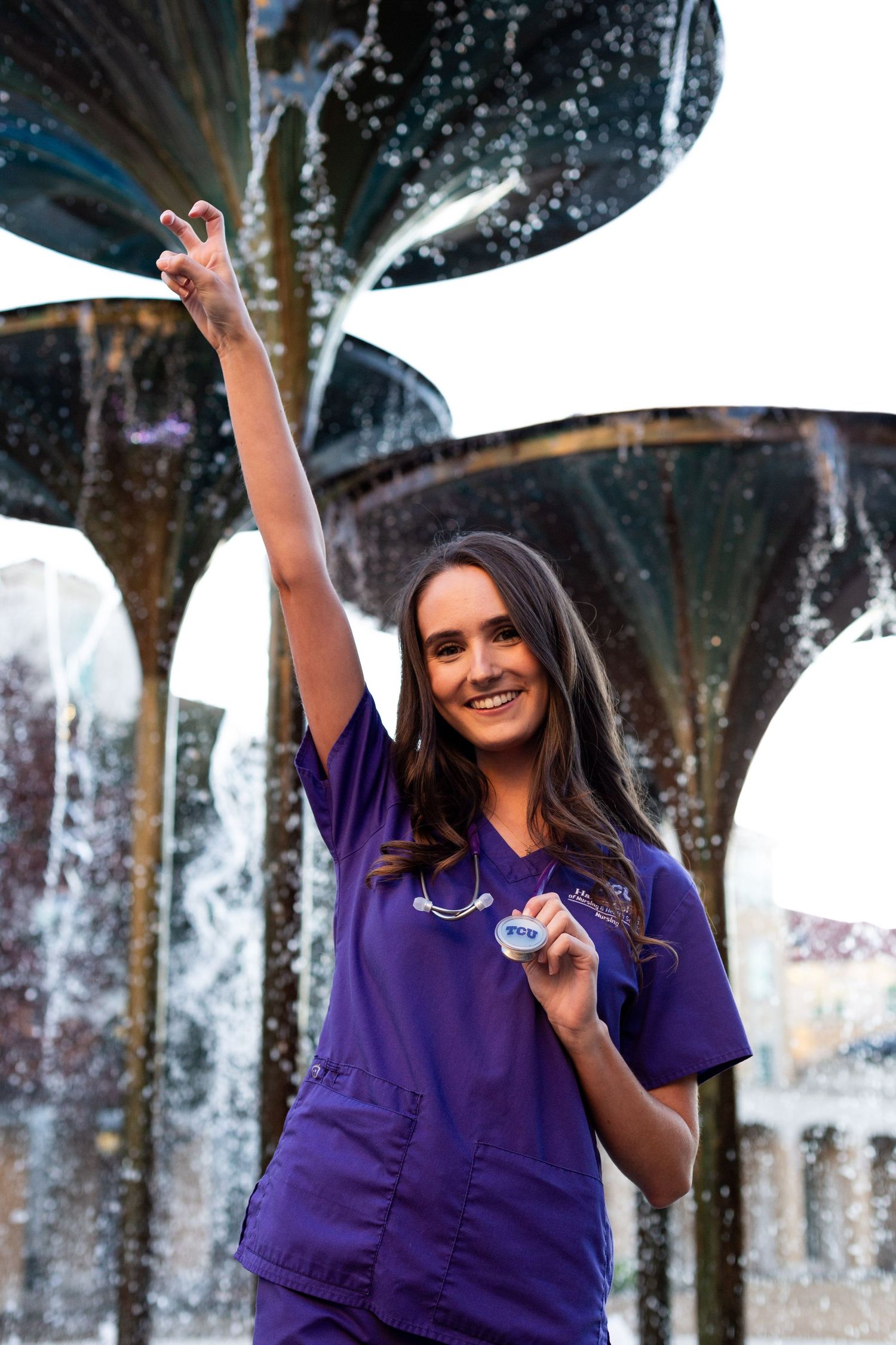 Healthcare worker in purple scrubs celebrates joyfully in front of a decorative water fountain feature.