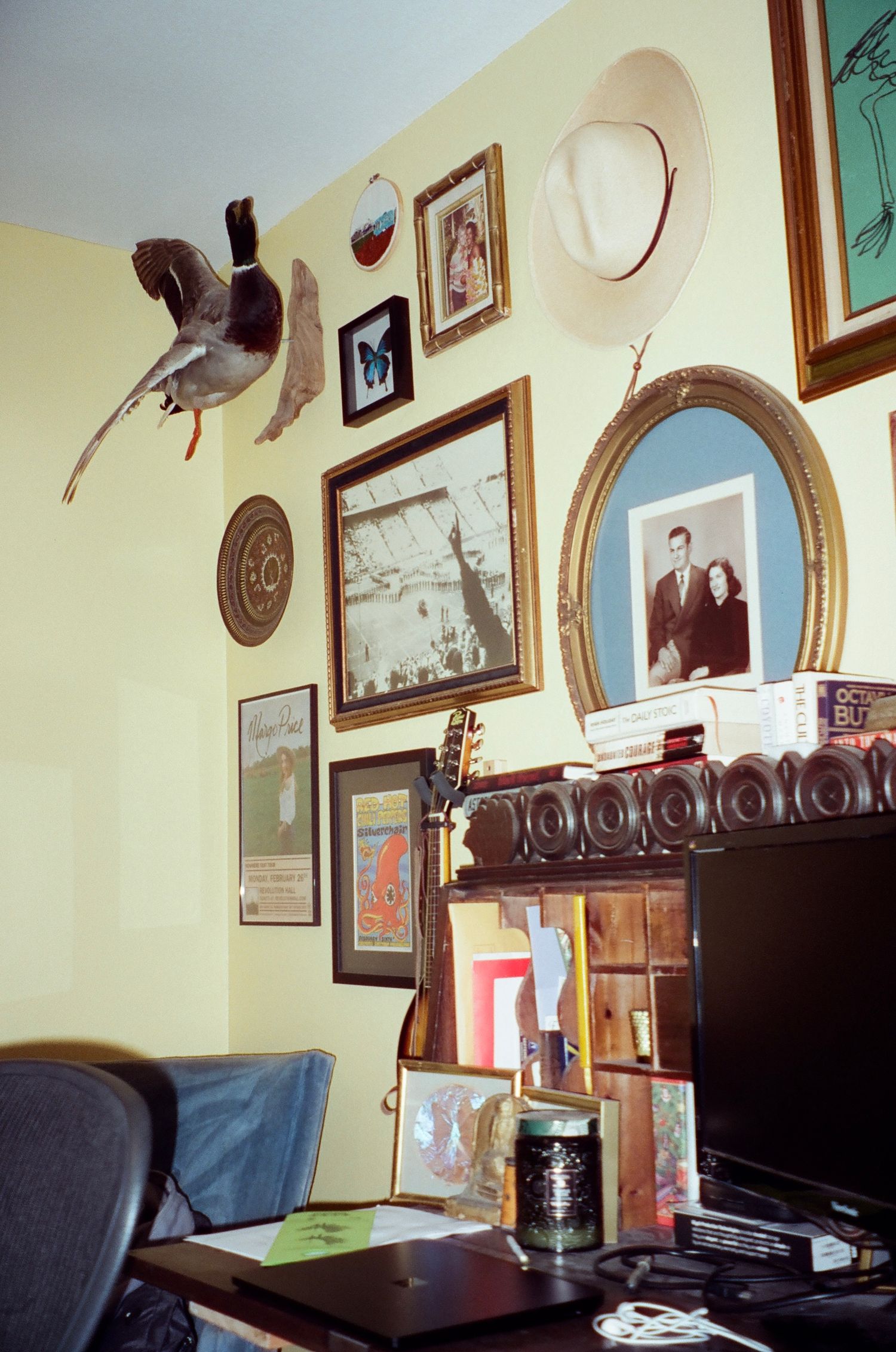 A cozy room corner with vintage framed photographs and artwork decorating pale yellow walls above a wooden desk setup.