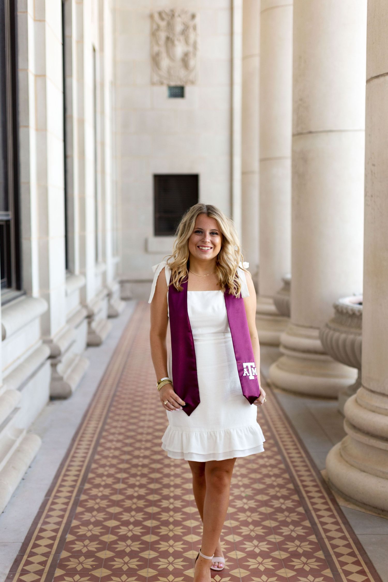 A white sleeveless dress with a purple blazer stands out against ornate columns and tiled floor in an elegant hallway.