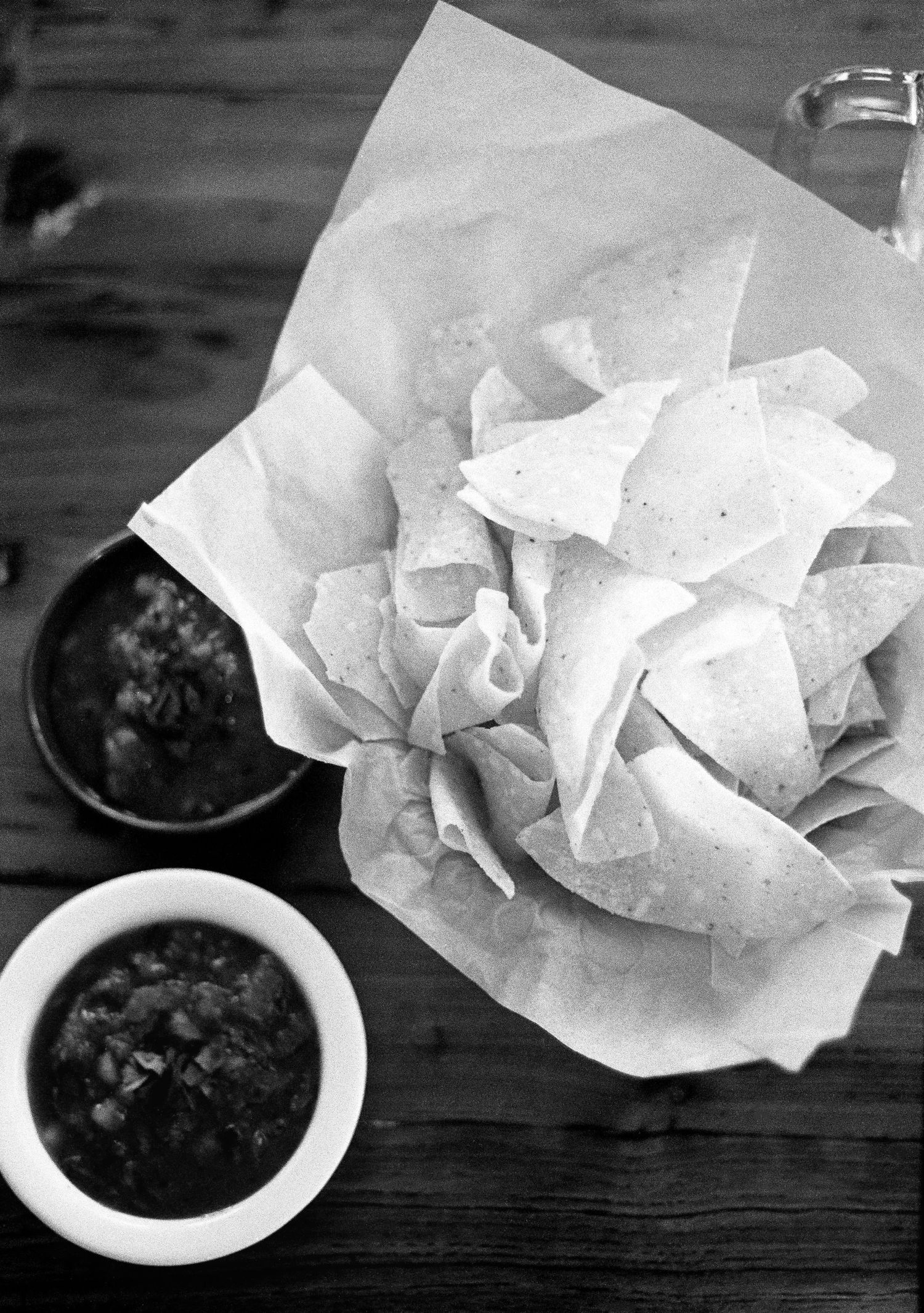 Black and white image of tortilla chips on white paper with small bowls of dark salsa on a wooden table.