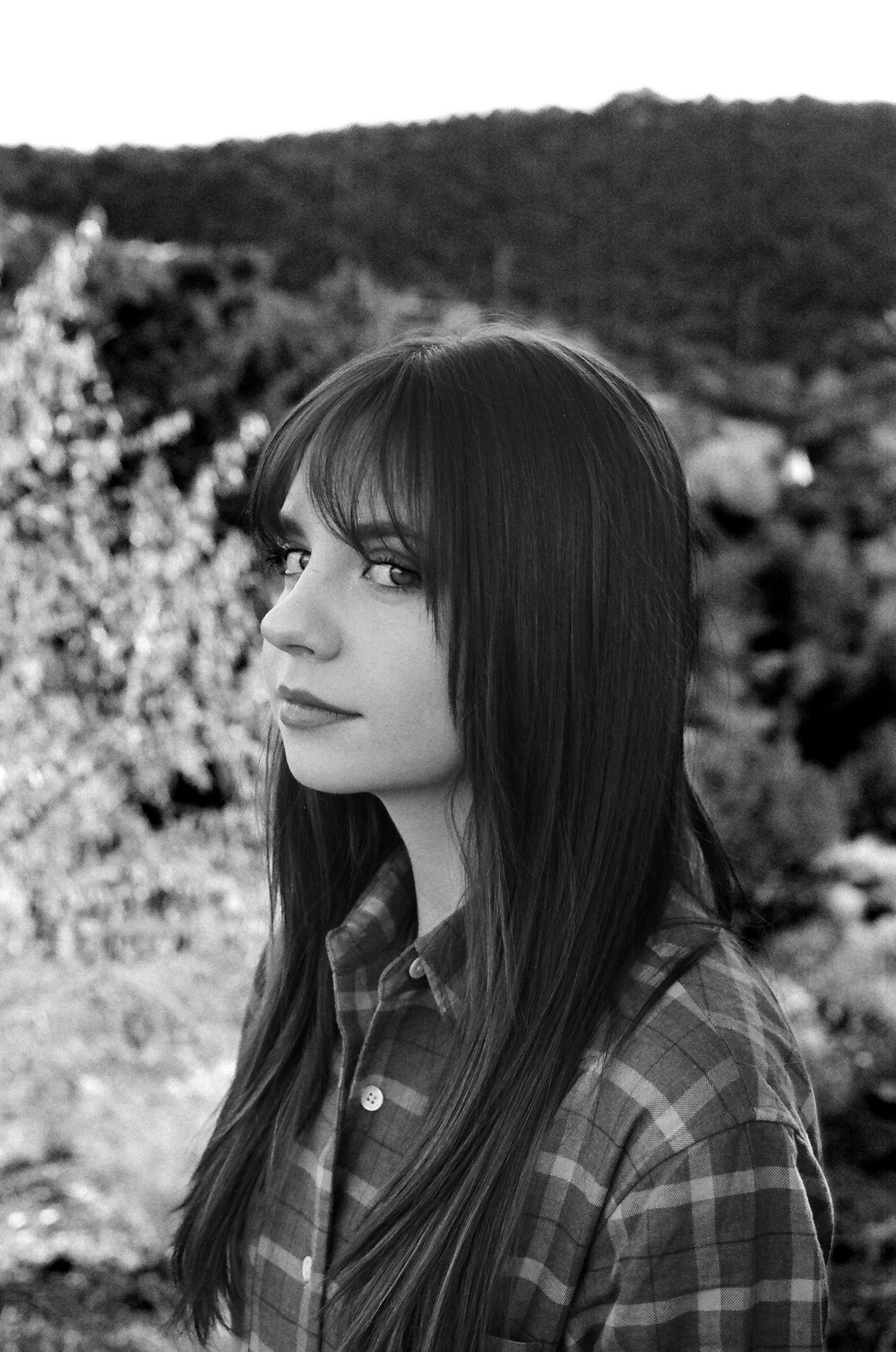 A black and white portrait of someone in a plaid shirt against a natural outdoor background.